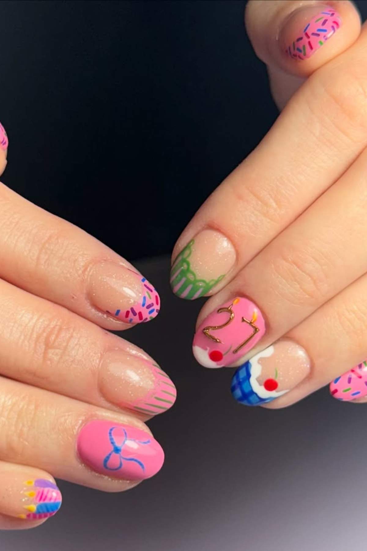 Hands with almond-shaped nails painted with colorful birthday-themed nail art, including sprinkles, cupcakes, a blue bow, and the number 27.