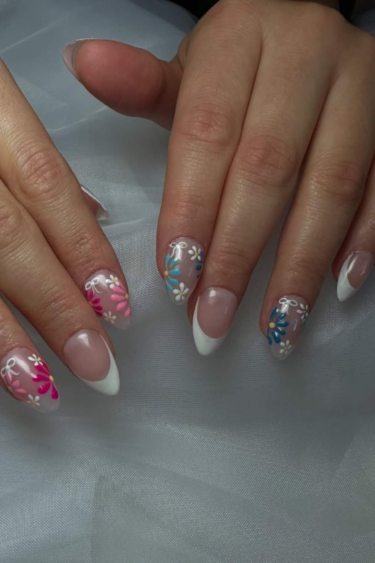 Hands with manicured nails featuring a French tip style and colorful floral designs in blue, pink, and white on a light sheer background.