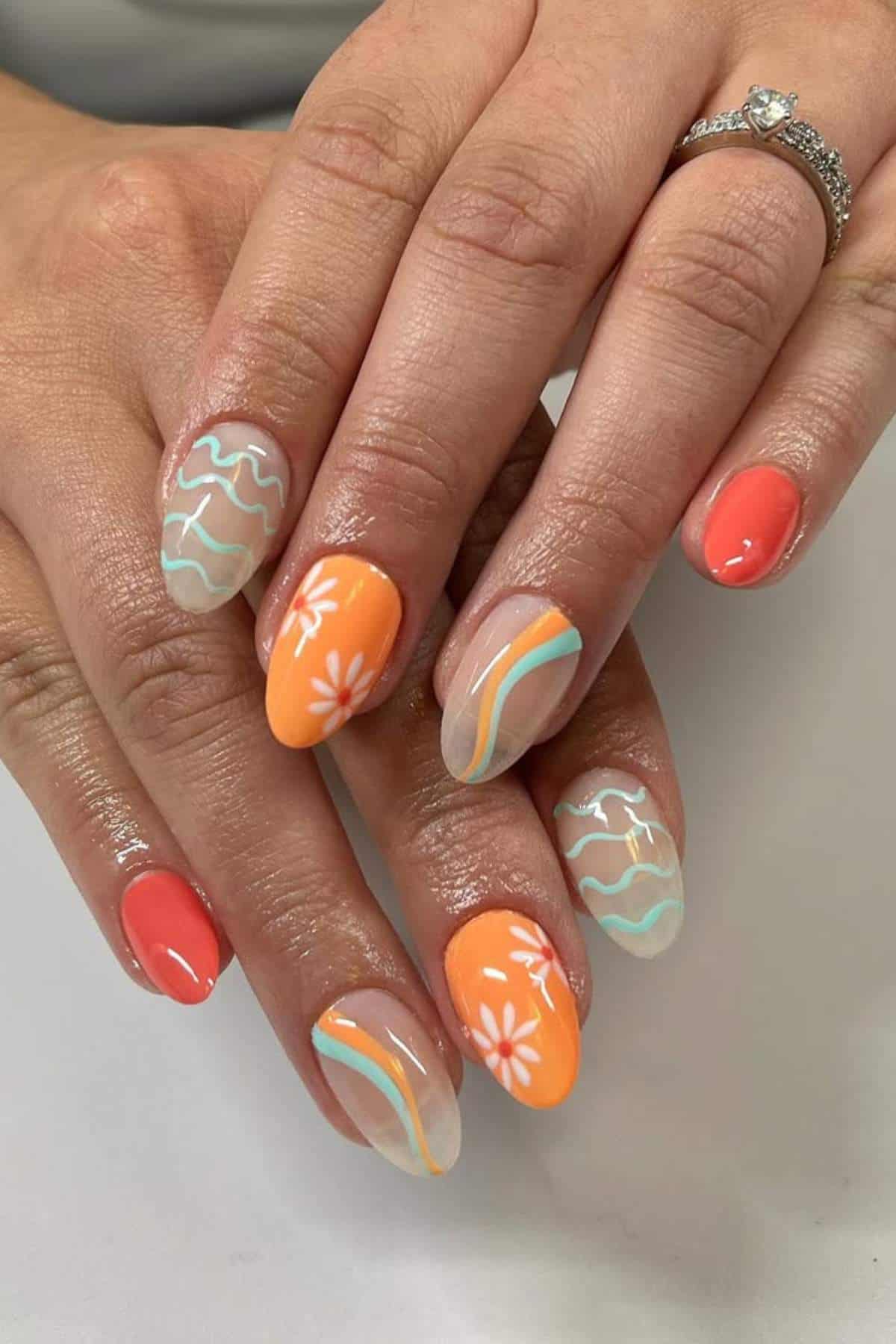 Hands with manicured nails featuring orange, coral, and nude polish, decorated with white floral designs, wavy lines, and a silver ring with a small gemstone.