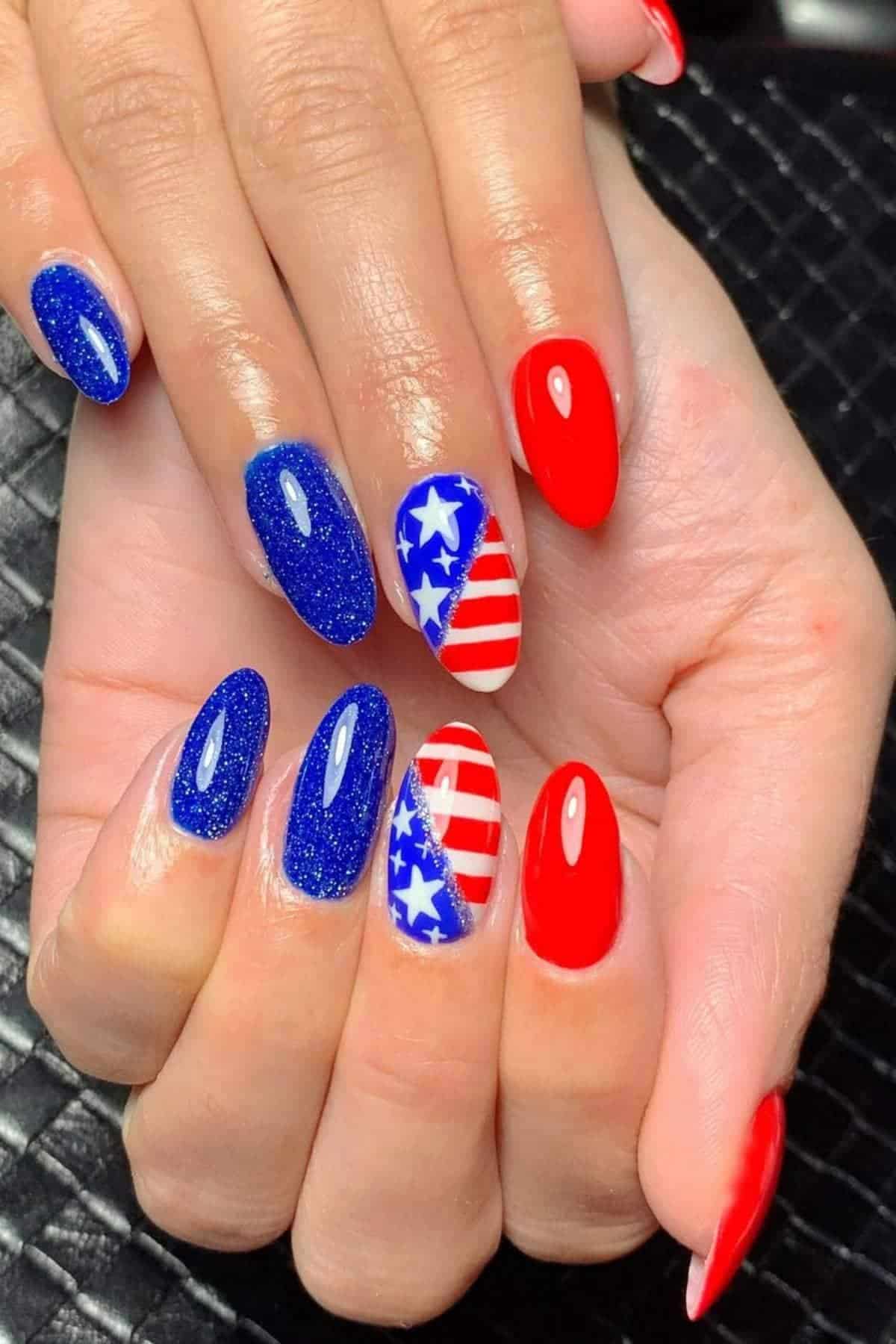 A hand with manicured nails features blue glitter polish, solid red polish, and accent nails with white stars and red stripes resembling the American flag.