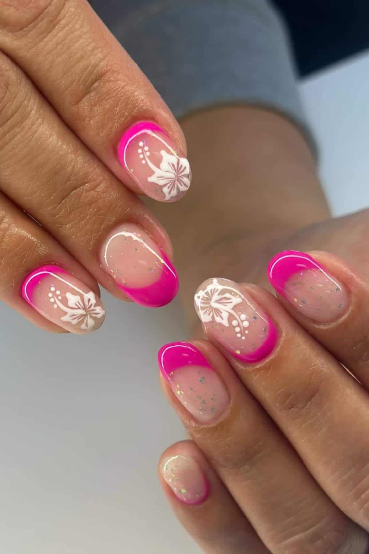 Hands with manicured nails featuring pink French tips, white floral designs, and subtle glitter on a nude base.