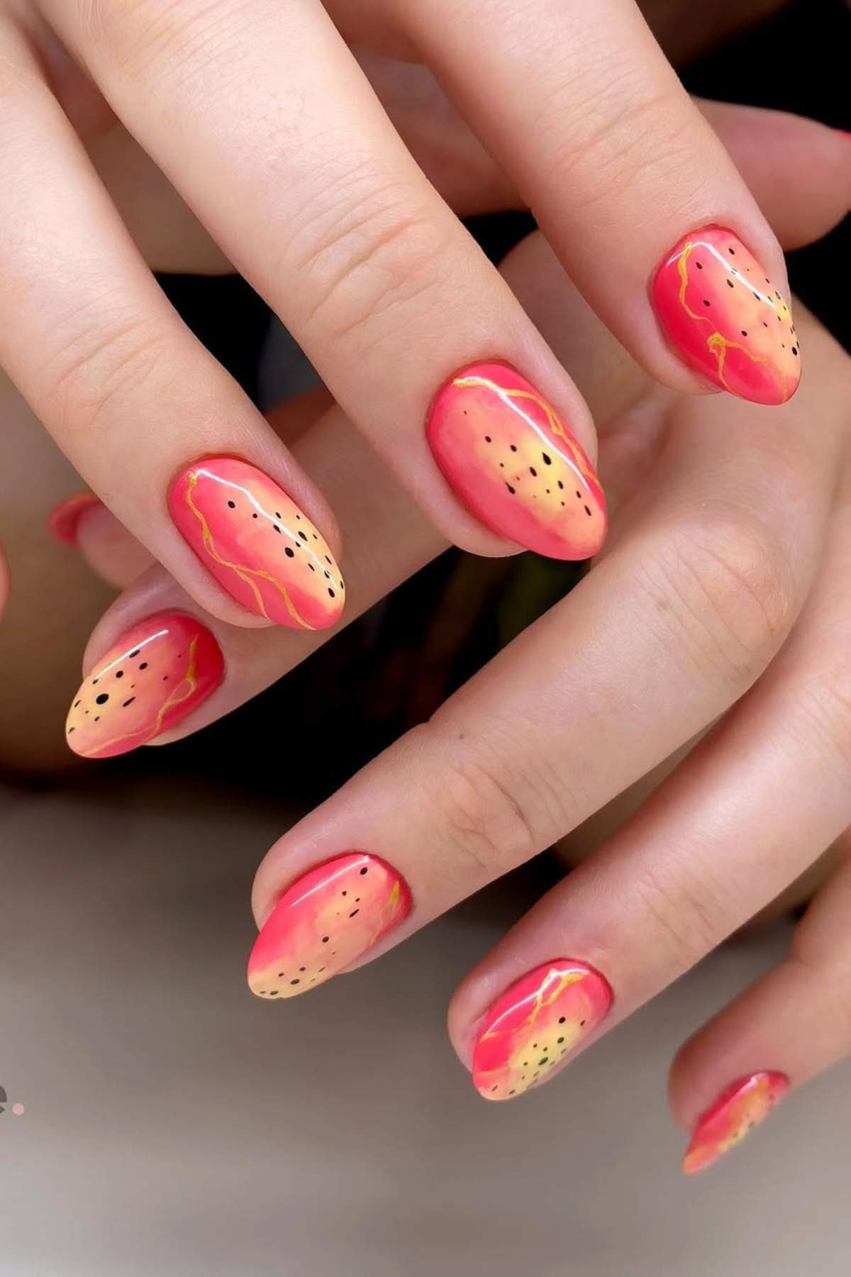 Fingernails with pink and yellow gradient polish, black dots, and gold accents resembling dragon fruit.