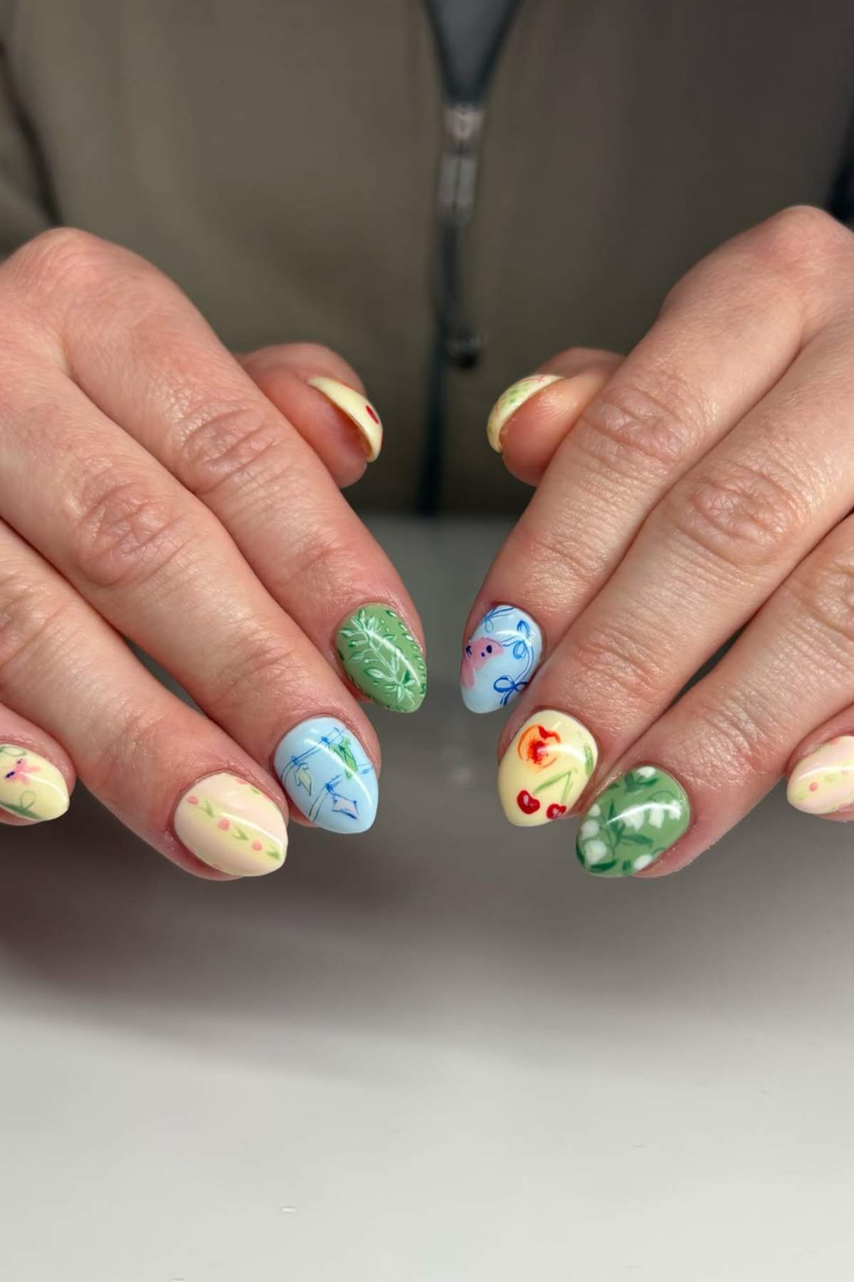 Hands with pastel-colored manicure featuring various hand-painted designs, including leaves, flowers, cherries, and geometric patterns on each nail.