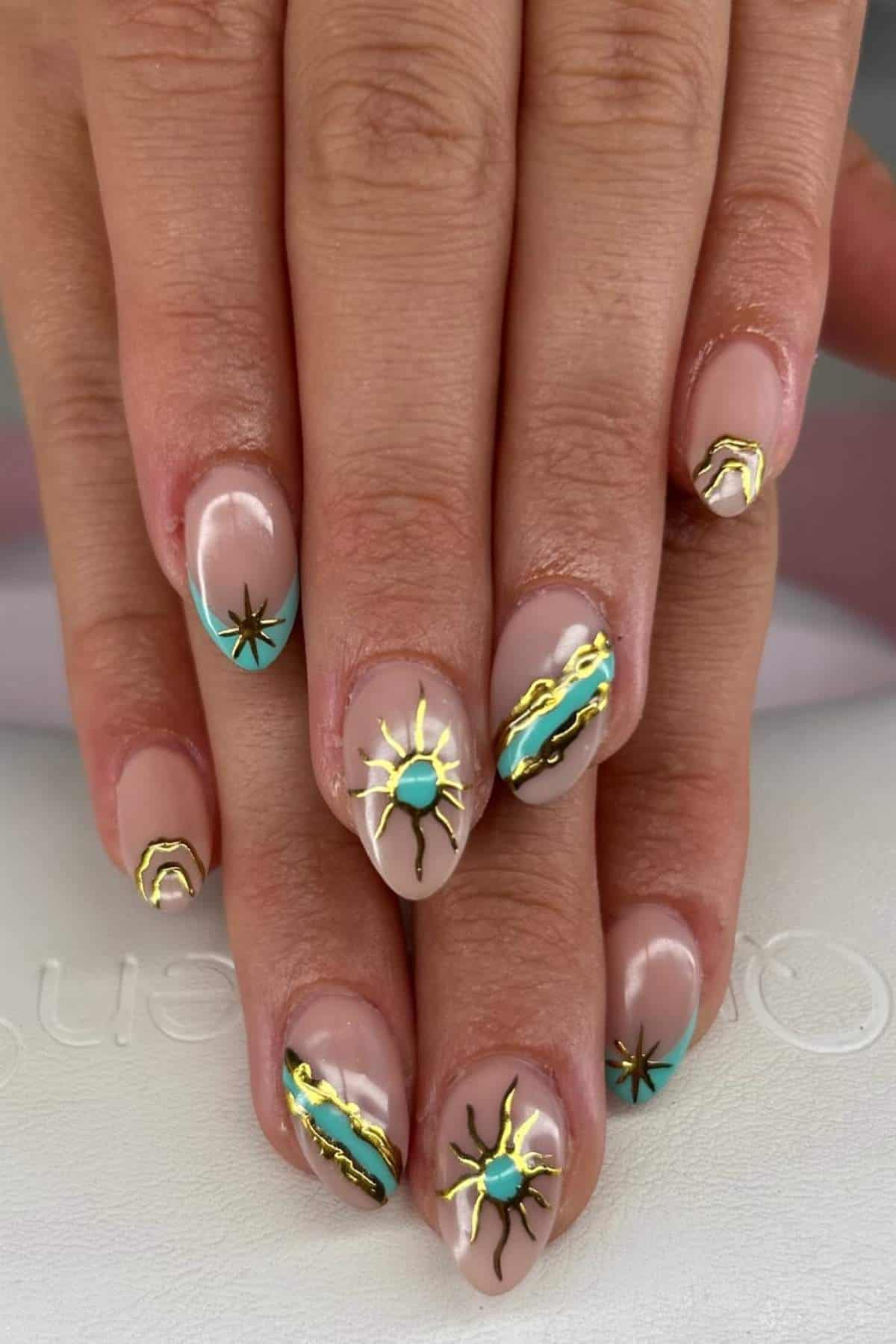 Hands with nude-colored nails featuring teal, gold, and black abstract sun and line designs.