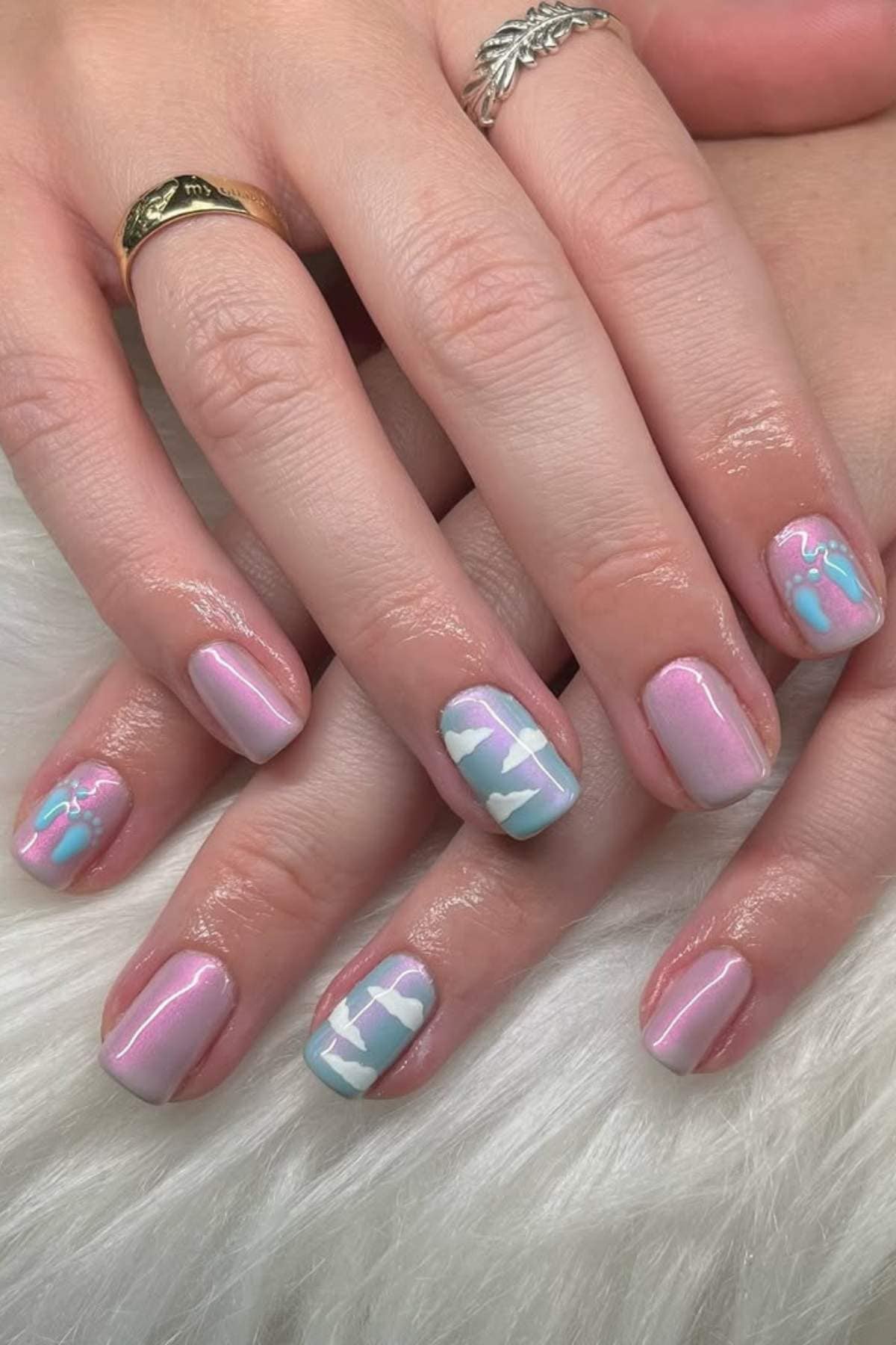 Hands with pastel pink and blue manicured nails featuring abstract and geometric designs, placed on a white furry surface.