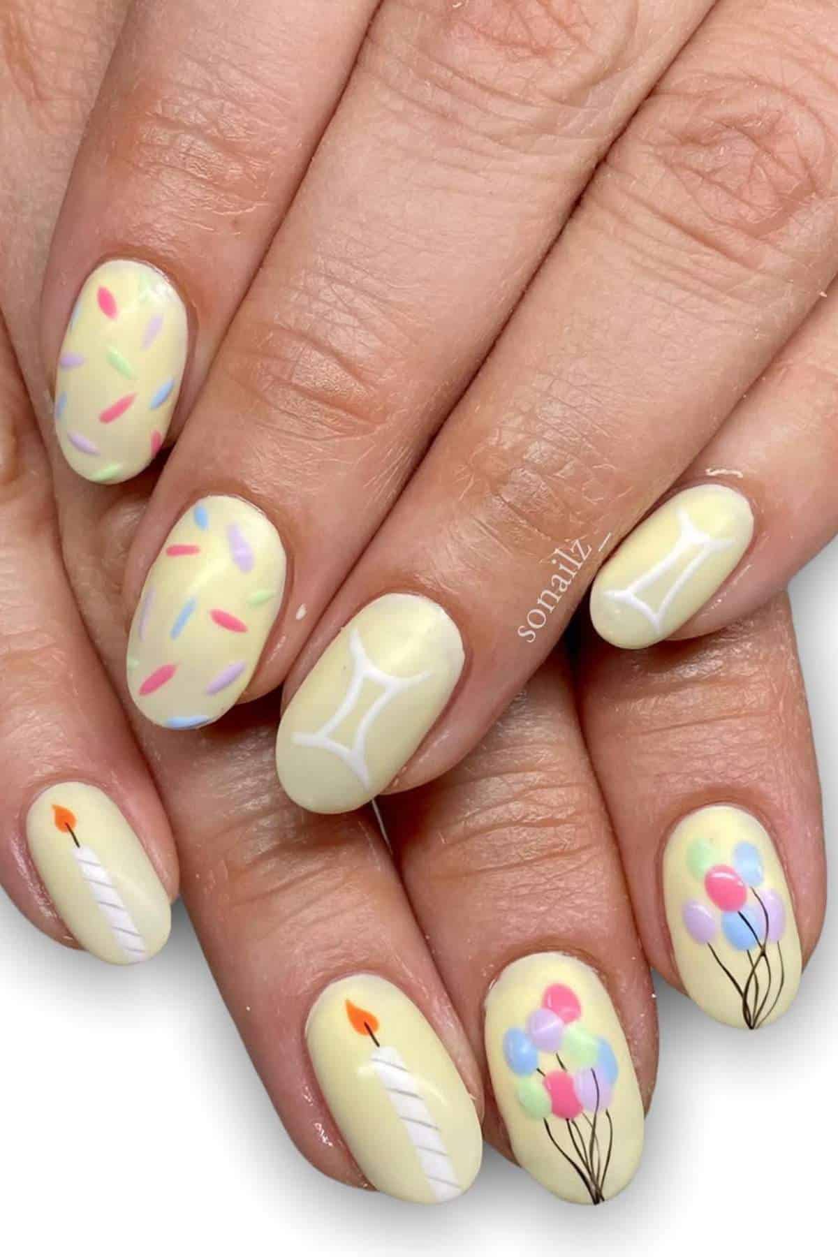 Hands with pastel yellow nail polish featuring birthday-themed designs: sprinkles, candles, and colorful balloons on each nail.