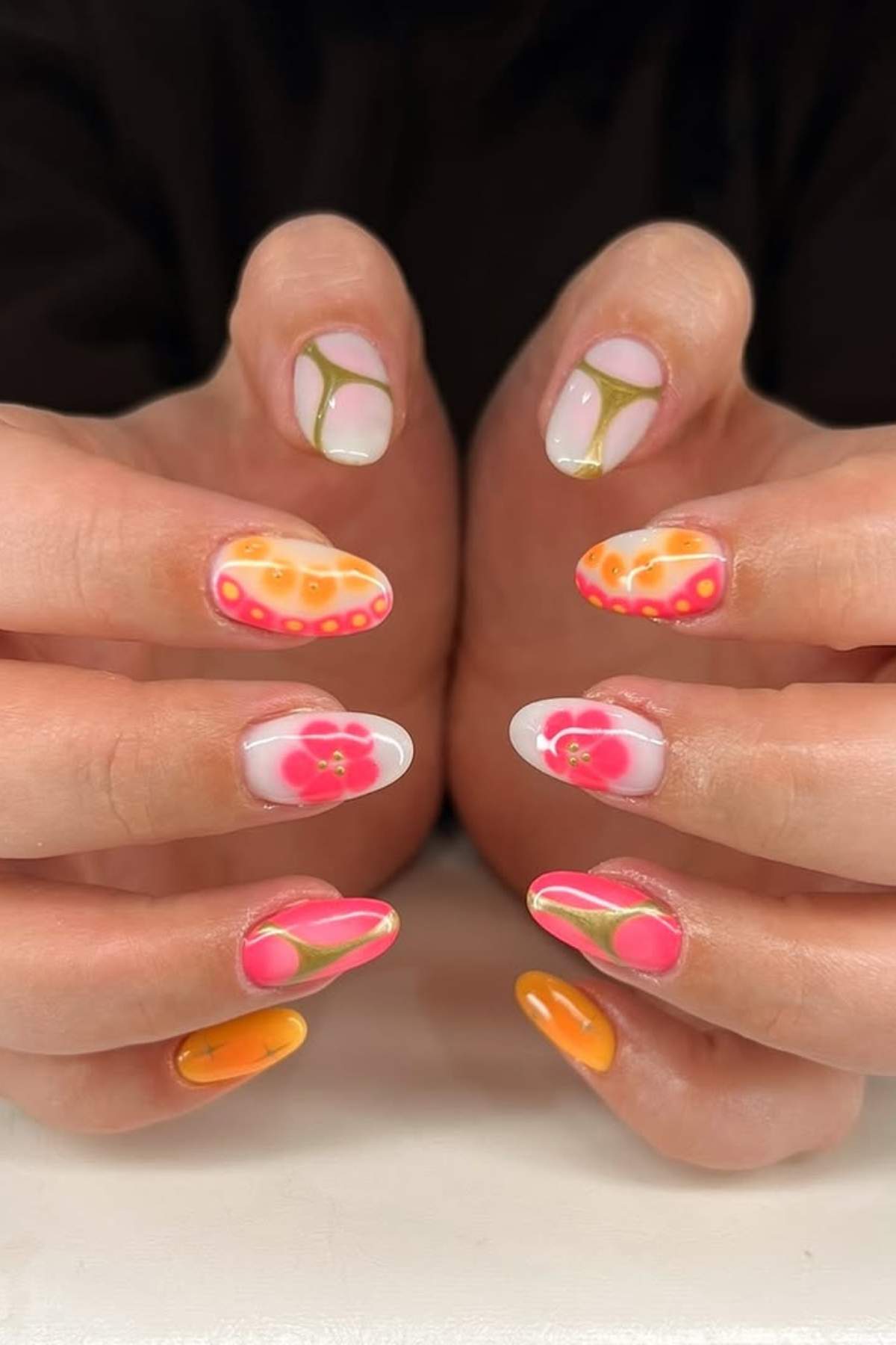 Hands with manicured nails featuring various colorful designs, including floral patterns, wavy lines, and a mix of pink, orange, and yellow tones.