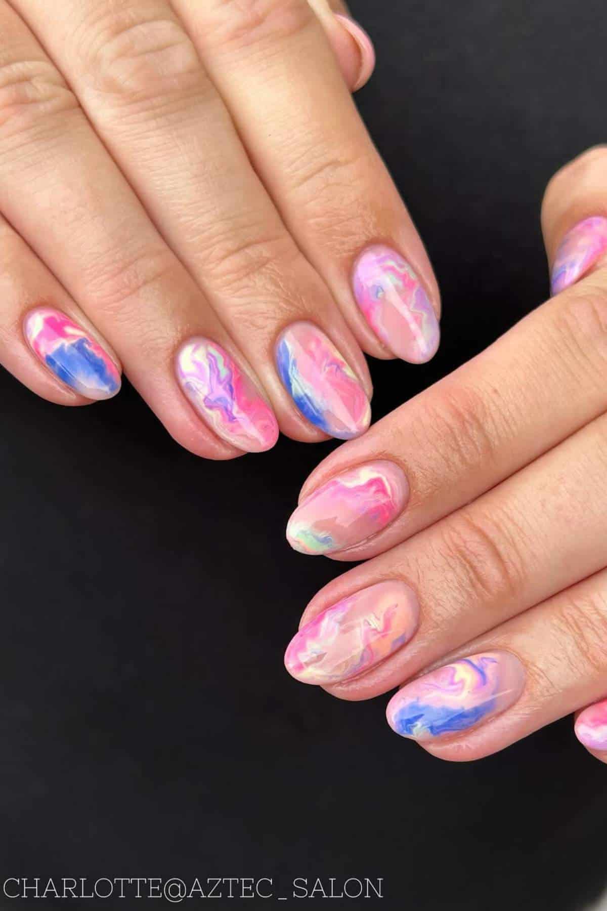 Close-up of hands with oval-shaped nails painted in a colorful marble design featuring pink, blue, and purple swirls on a black background.