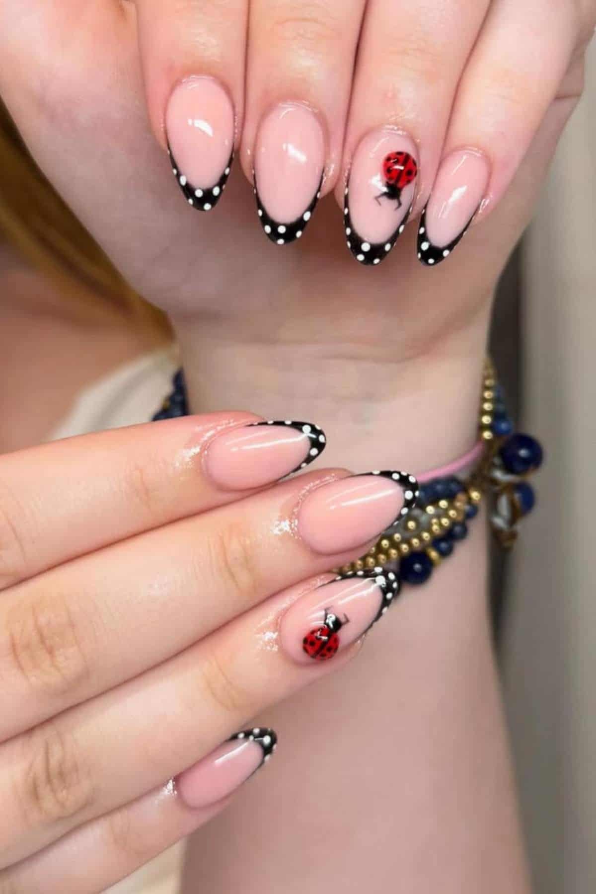 Hands with almond-shaped nails featuring a nude base, black French tips with white polka dots, and ladybug nail art on two accent nails.