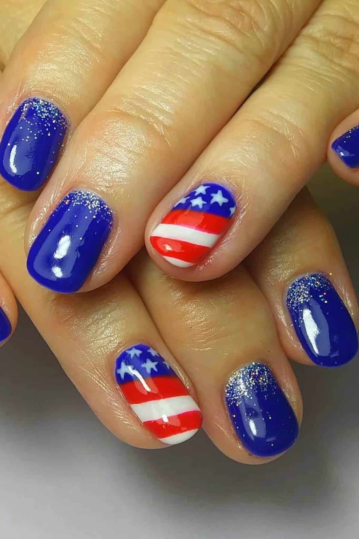 Close-up of fingernails painted blue with silver glitter; two accent nails feature a red, white, and blue stars and stripes American flag design.