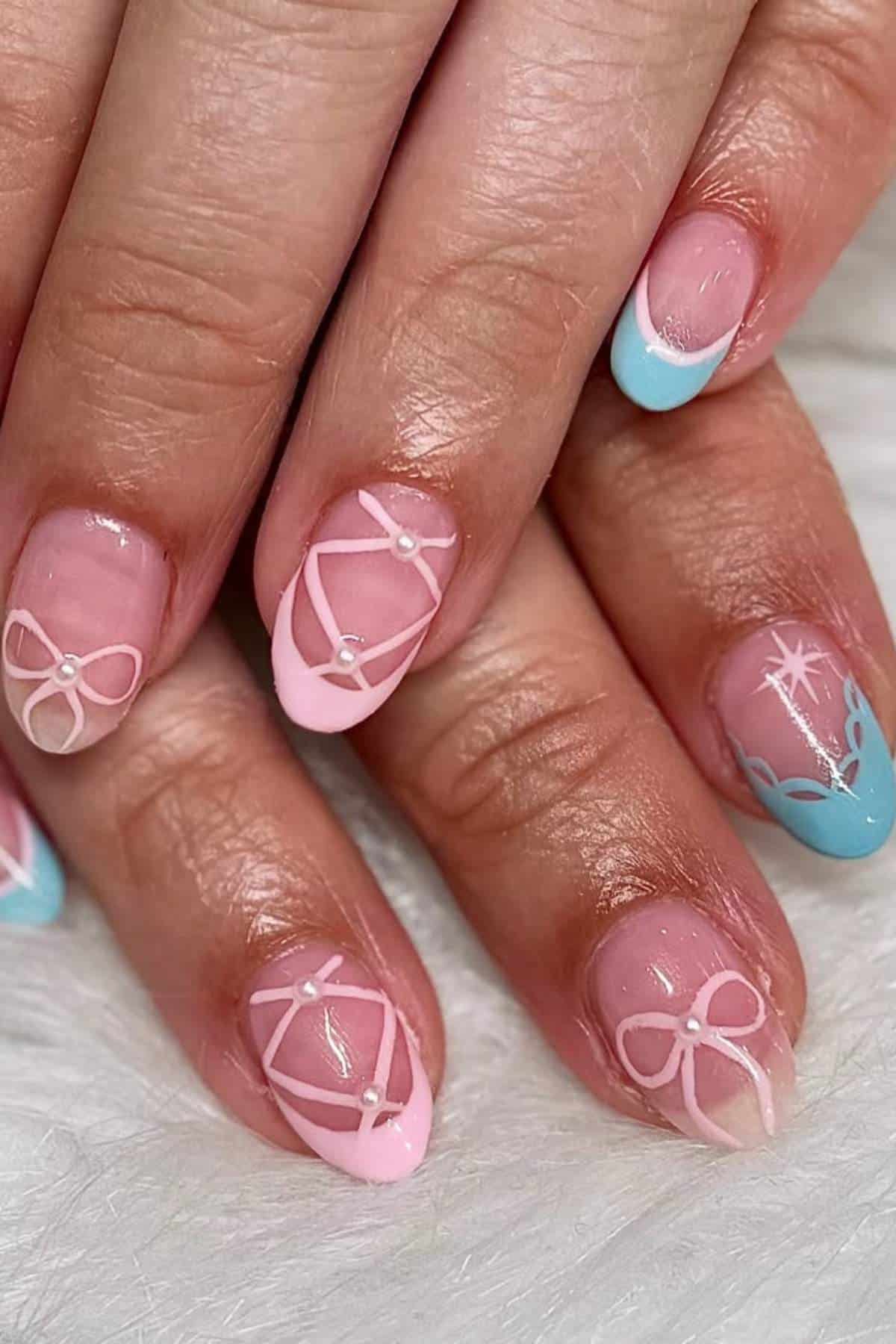 Close-up of hands with manicure featuring pink and blue tips, white bow designs, a small gem accent, and a white starburst pattern on one nail.