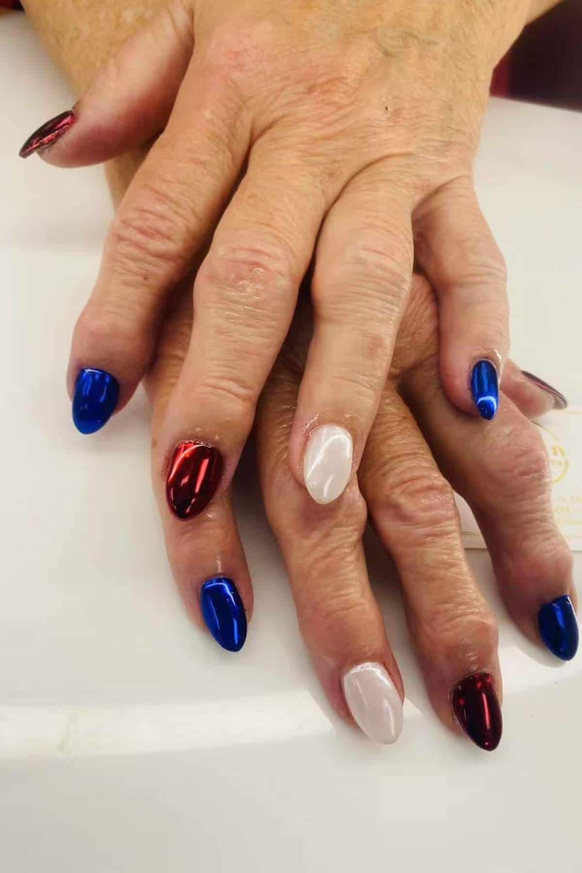 A pair of hands with manicured nails painted in metallic red, blue, and white colors, displayed on a white surface.