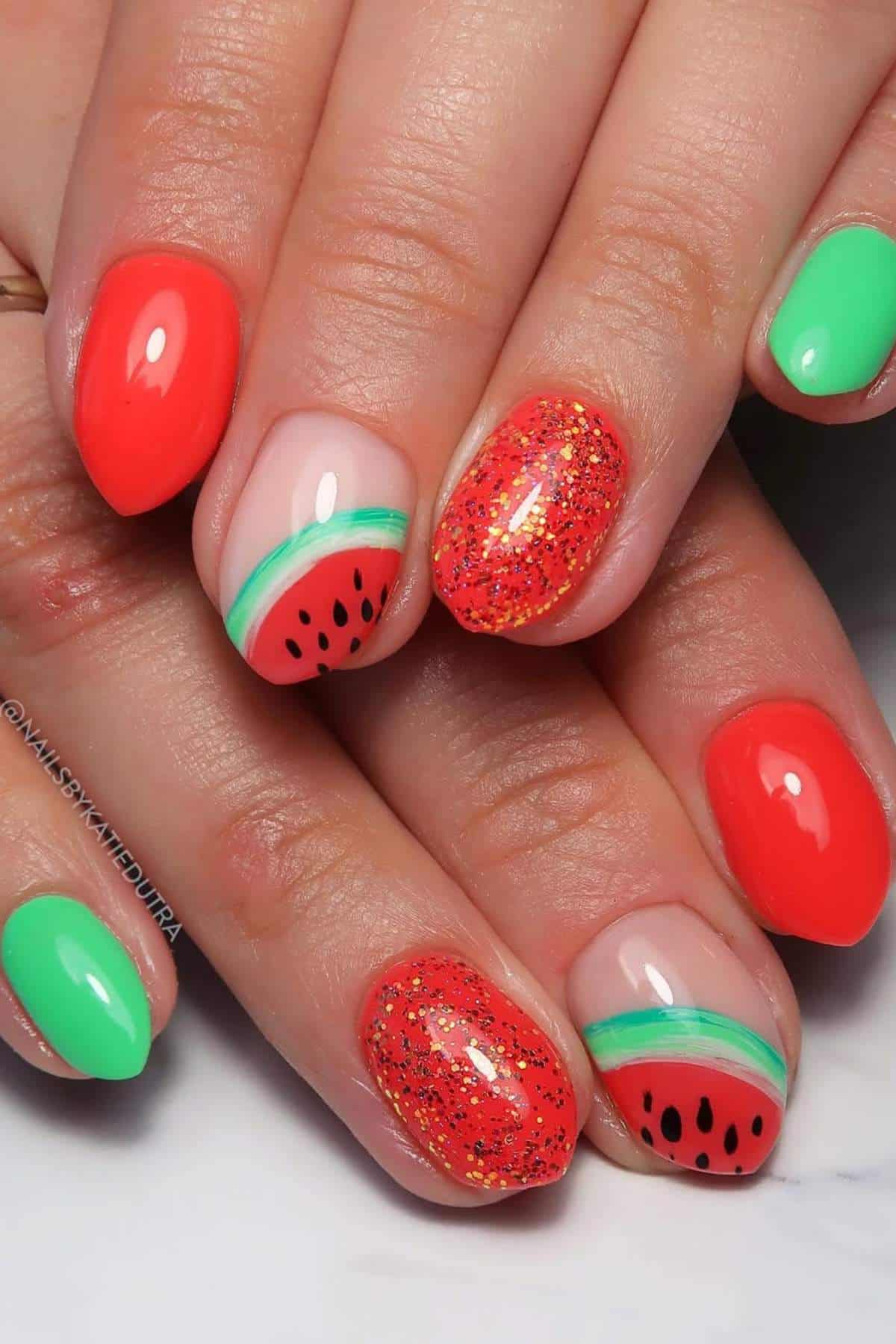 A hand with nails painted in a watermelon-inspired design, featuring red, green, glitter, and black seed-like details.