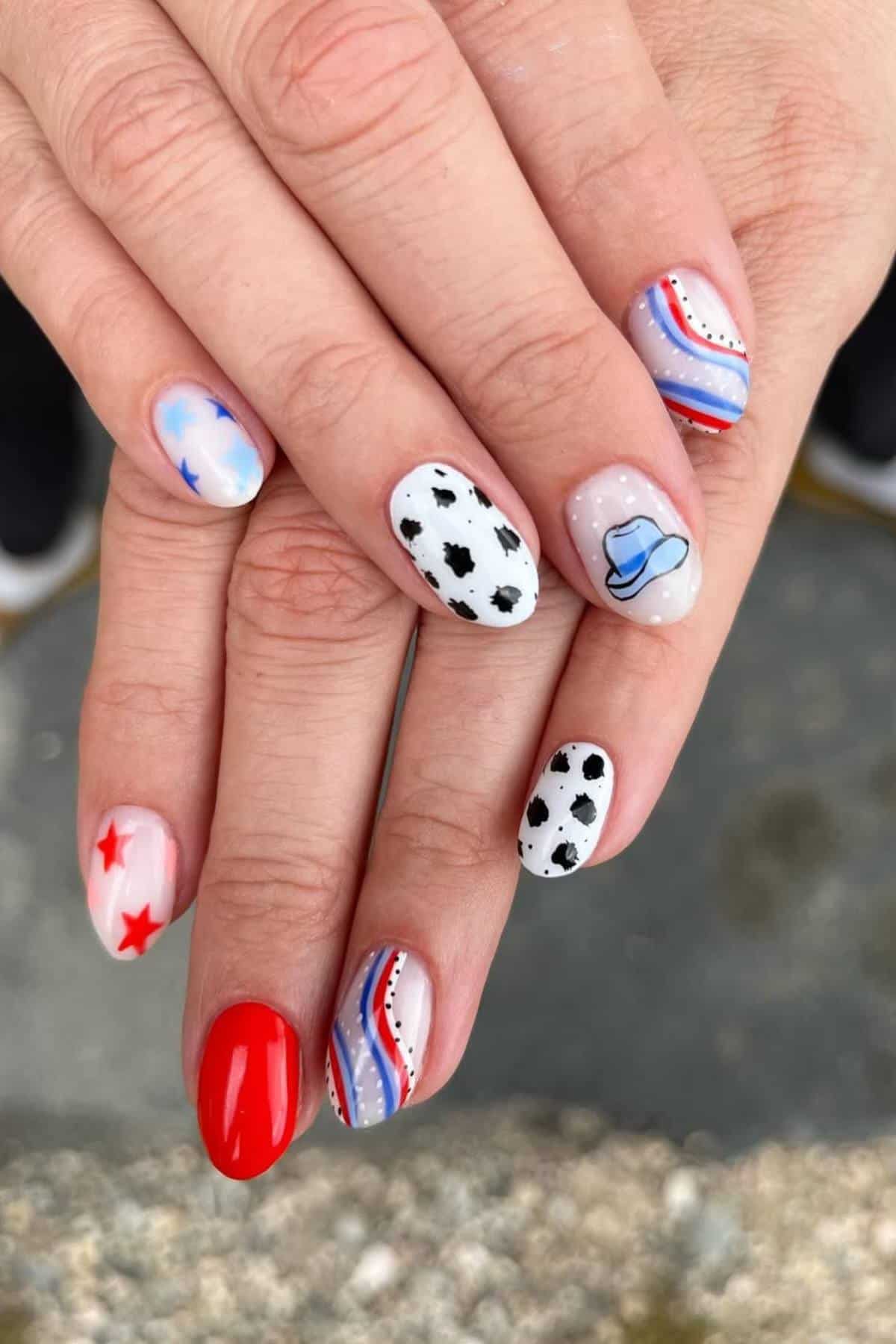 Hands with manicured nails featuring various designs including red stars, a cowboy hat, black cow spots, blue stars, red, white, and blue stripes, and solid red polish.
