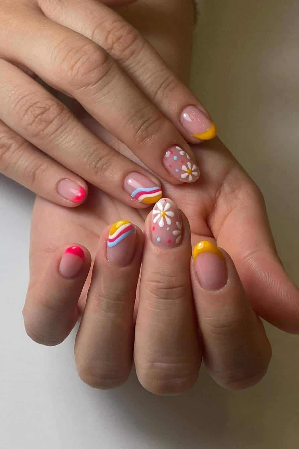 Hands with manicured nails featuring colorful designs, including flowers, polka dots, rainbow stripes, and yellow and pink French tips on a neutral base.