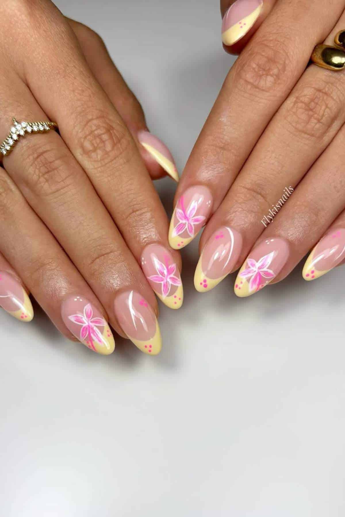 Hands with almond-shaped nails featuring yellow French tips, pink floral designs, and small polka dots. Person is wearing gold and diamond rings.