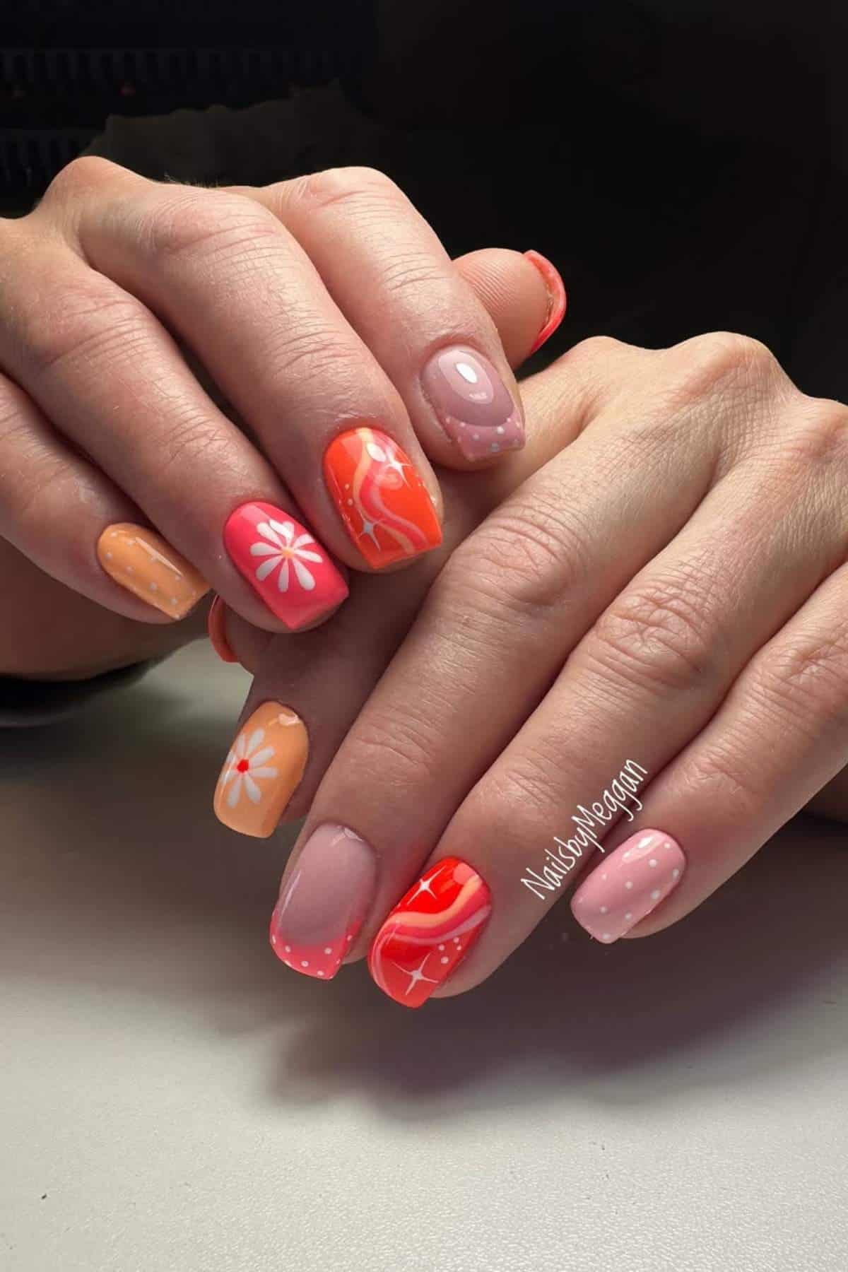 Close-up of hands with manicured nails featuring orange and pink shades, floral designs, polka dots, and abstract patterns.