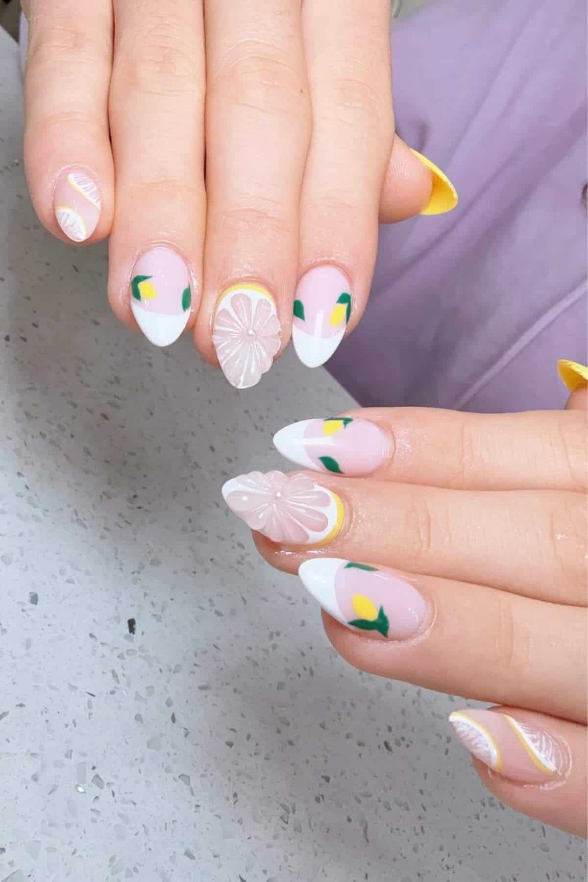 Hands displaying almond-shaped nails with pastel pink polish, white tips, yellow and green lemon designs, and raised floral accents on two nails, resting on a speckled surface.