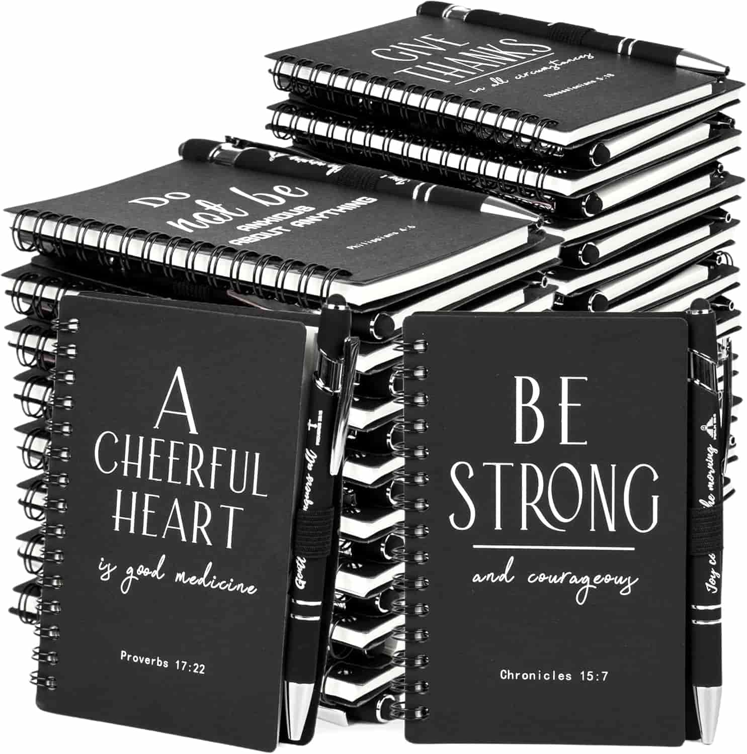 A stack of black spiral notebooks with motivational Bible verses on the covers, each notebook paired with a matching pen.