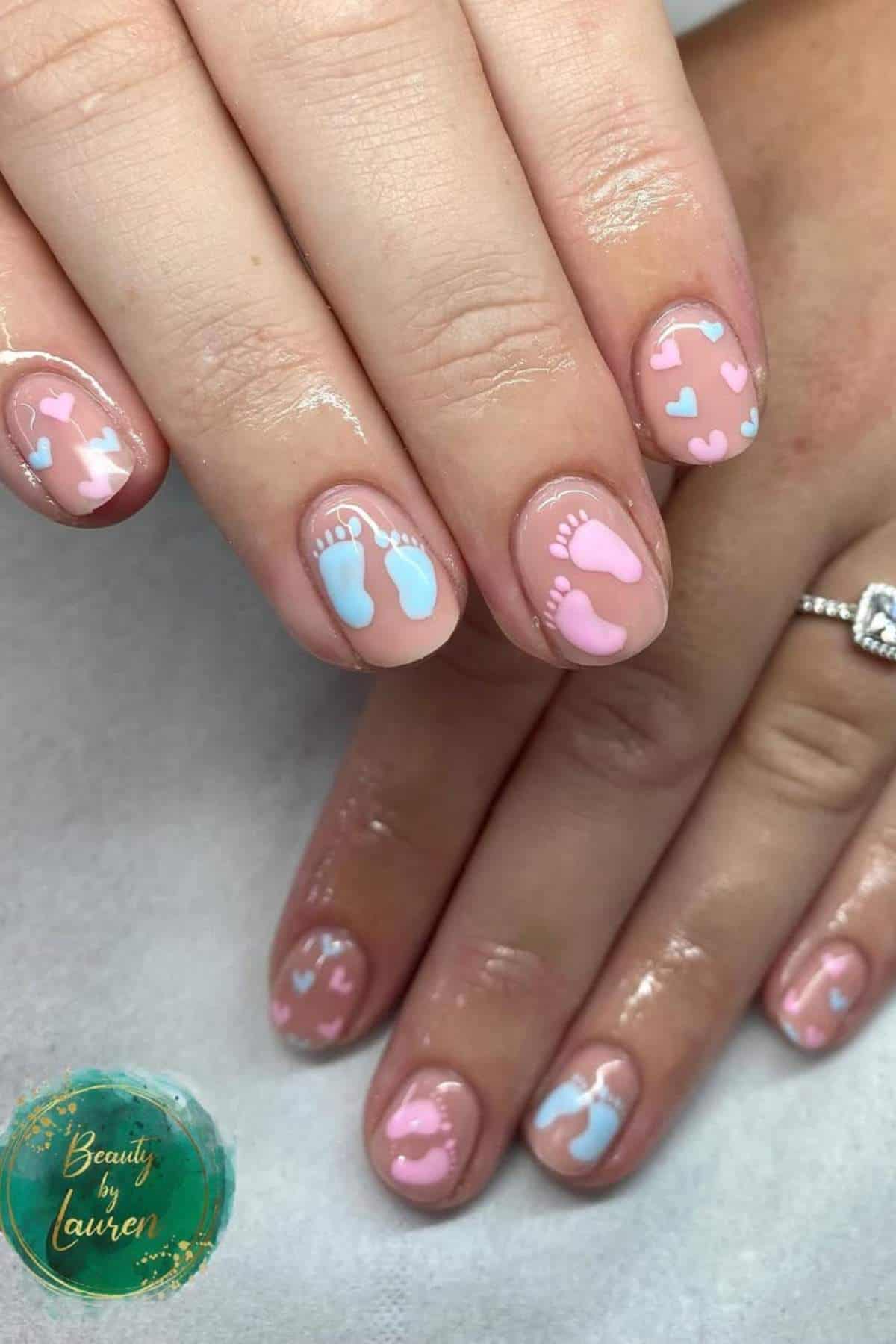 Hands with manicured nails featuring pink and blue baby footprints and small heart designs on a nude base.