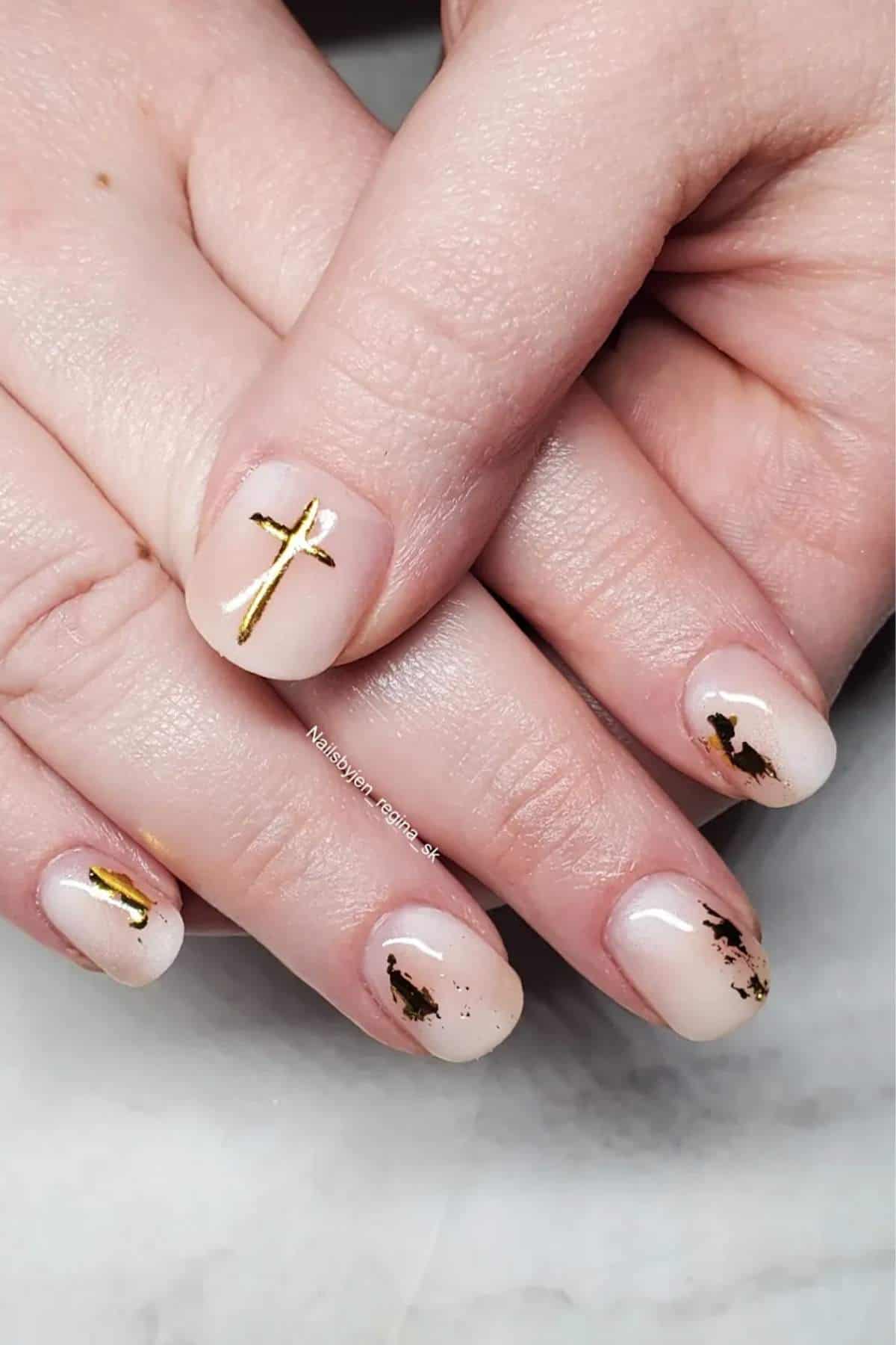 Hands with short, nude-colored nails featuring gold cross designs and abstract gold accents on a marble surface.