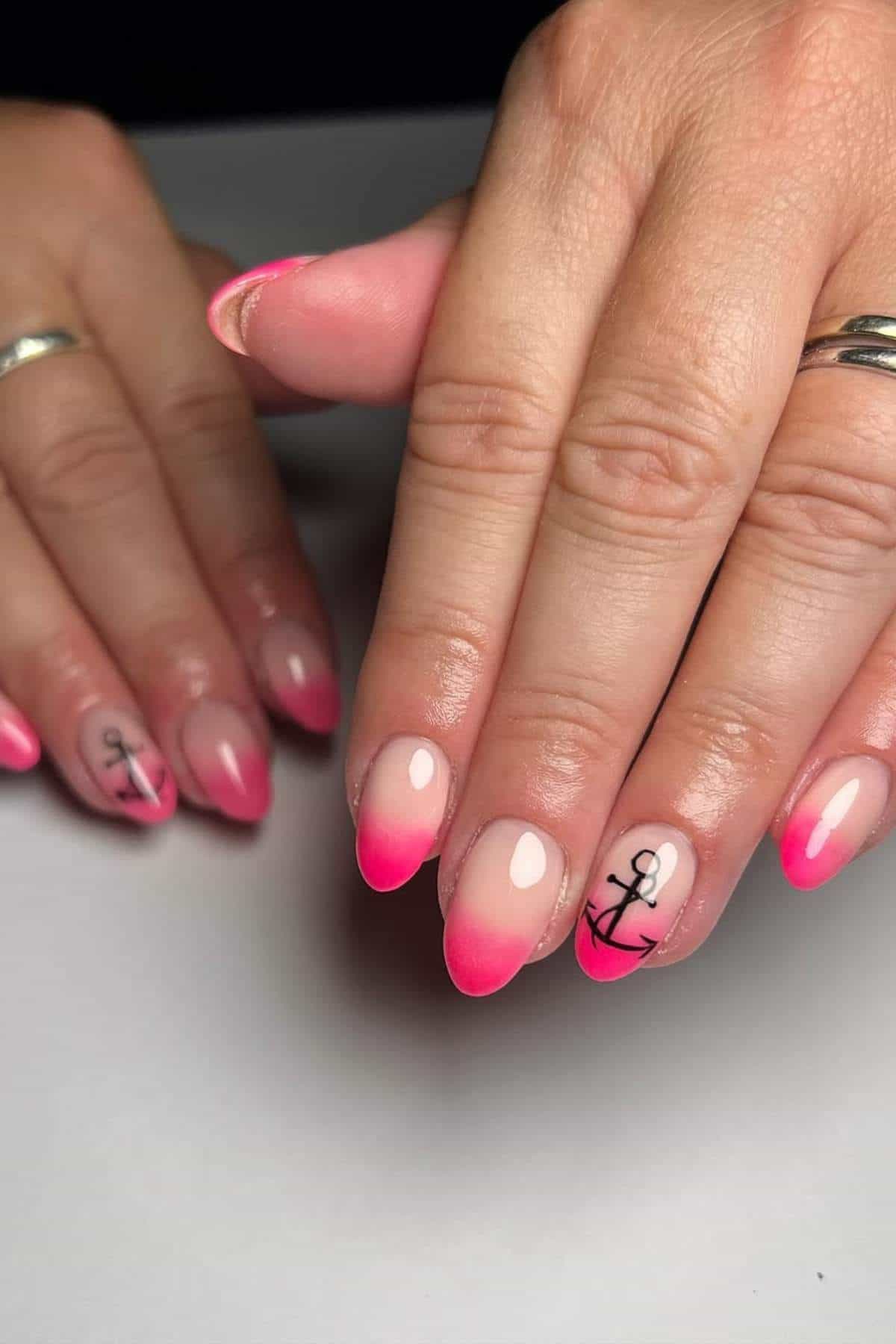 Hands with almond-shaped nails featuring pink ombre tips and black anchor designs on two accent nails.