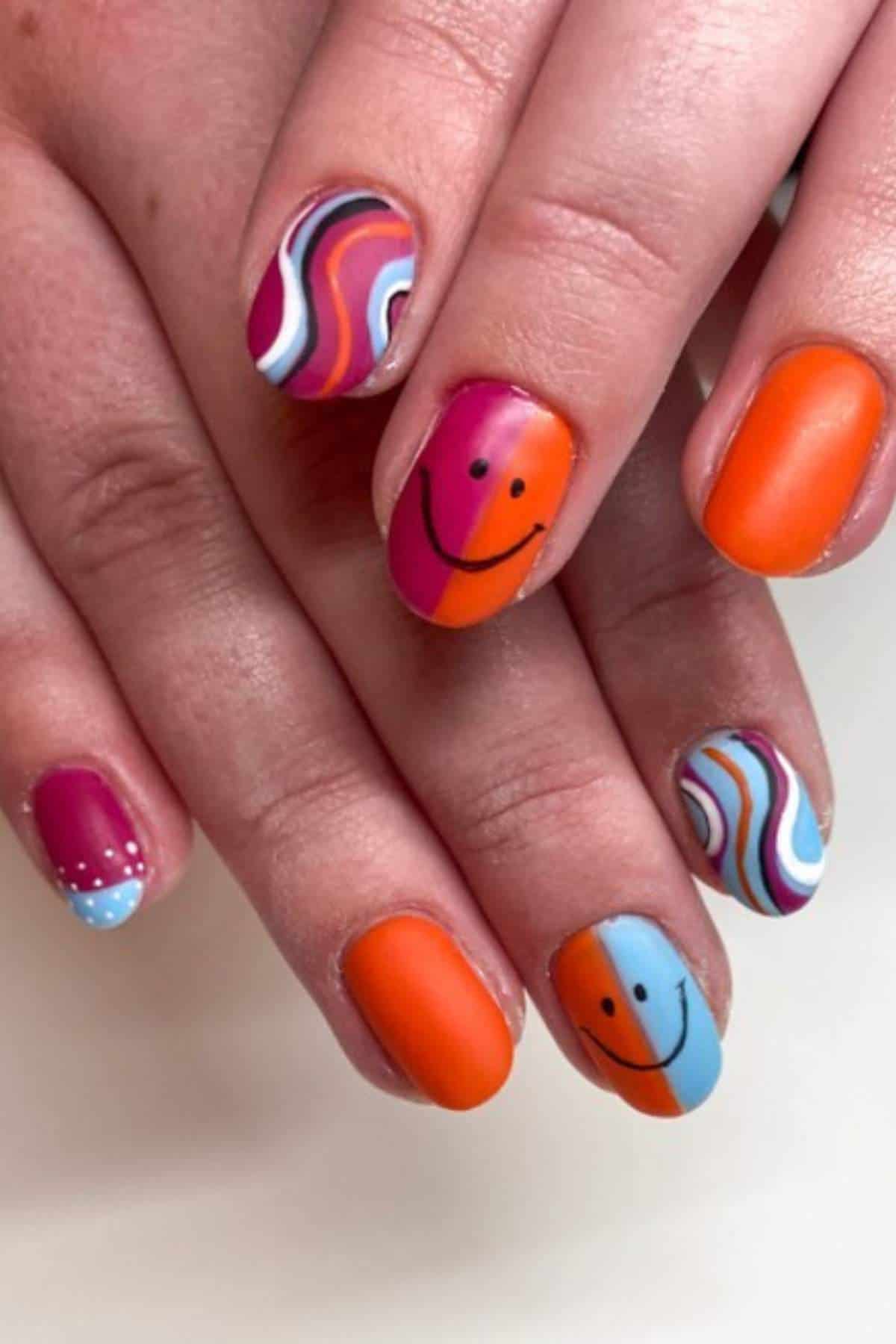Hands with manicured nails featuring orange, blue, and magenta matte polish, smiley face designs, wavy patterns, and one nail with small white dots.