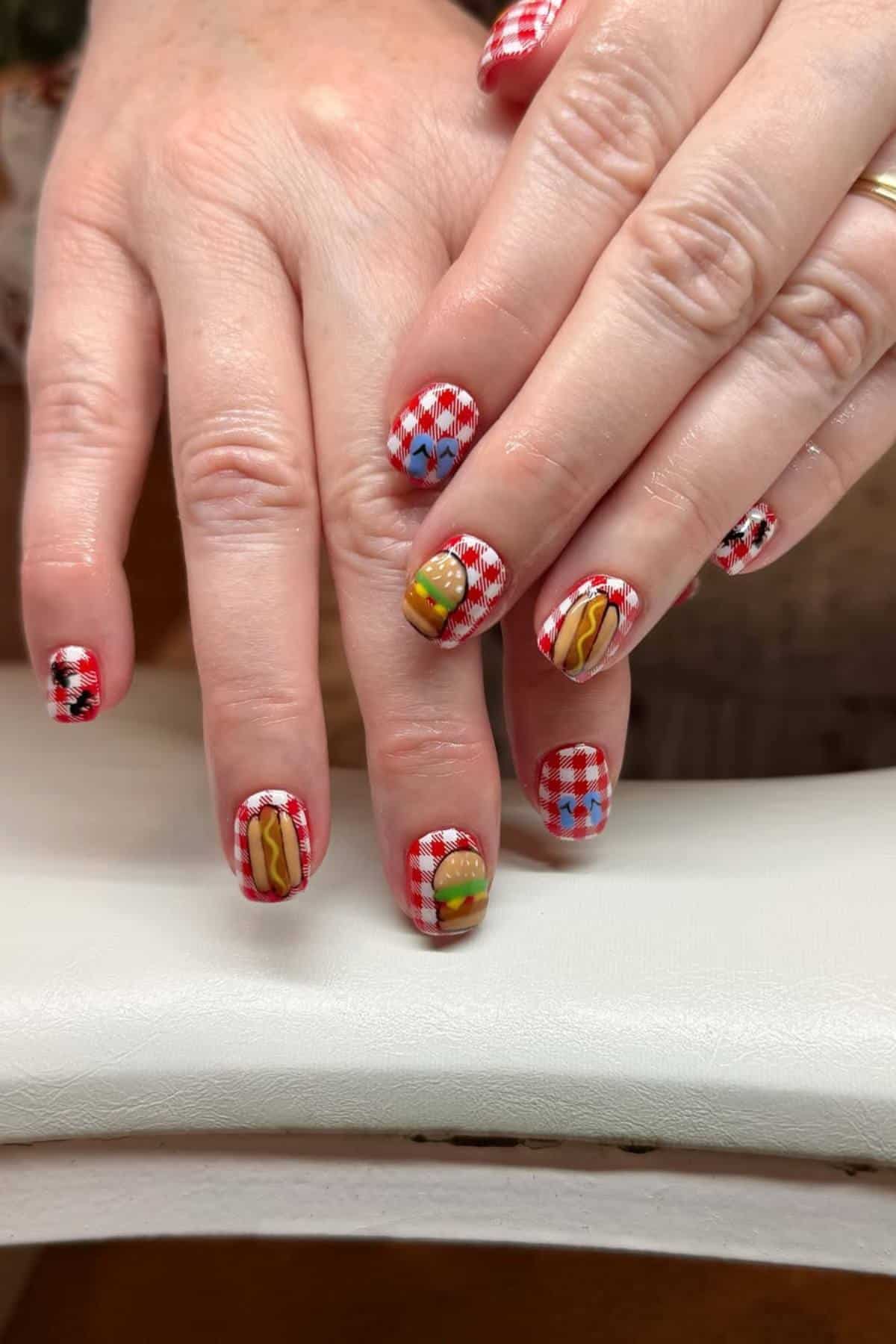 Hands with red and white checkered nail art, featuring designs of a hamburger, hotdog, and condiments on some fingernails.