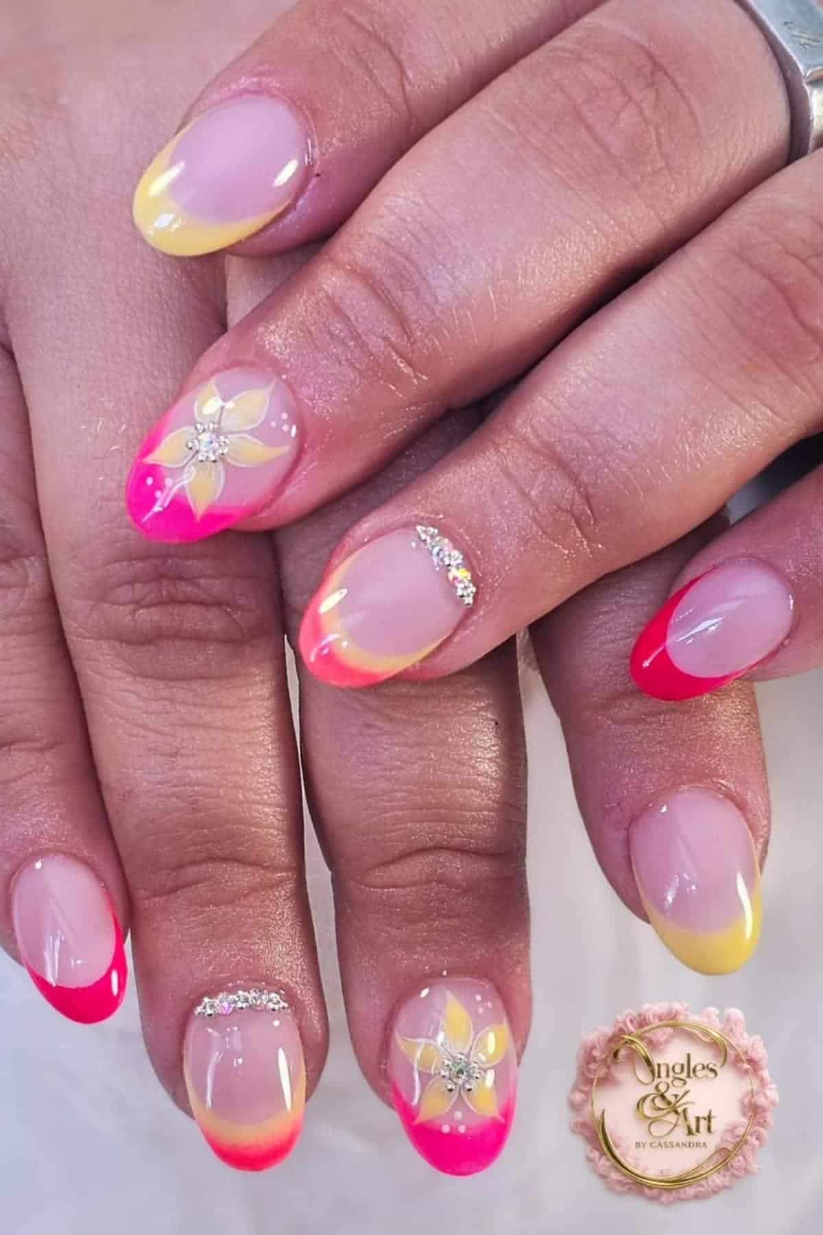 Hands with almond-shaped nails featuring pink and yellow French tips, rhinestone accents, and a floral design on two nails with a salon logo in the corner.