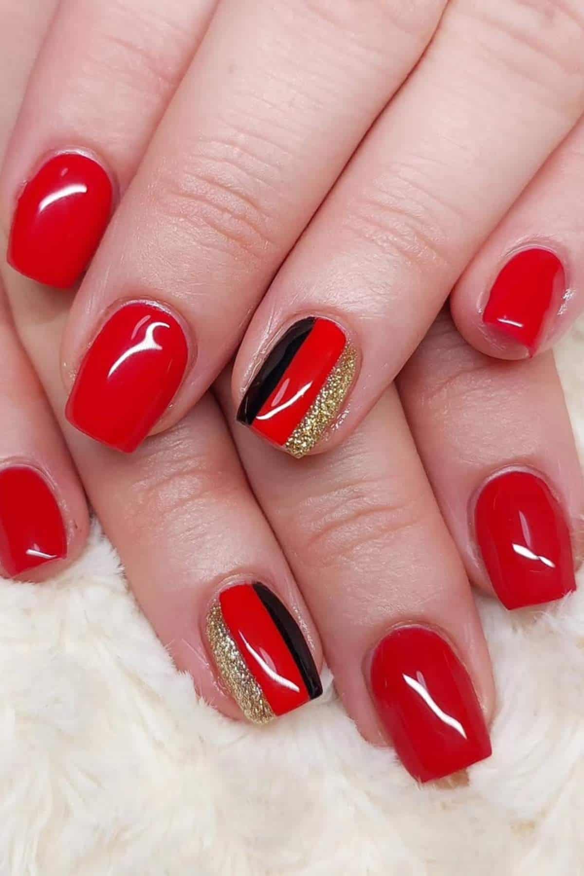 Close-up of hands with red nail polish; ring fingers feature gold glitter and a diagonal black accent design.