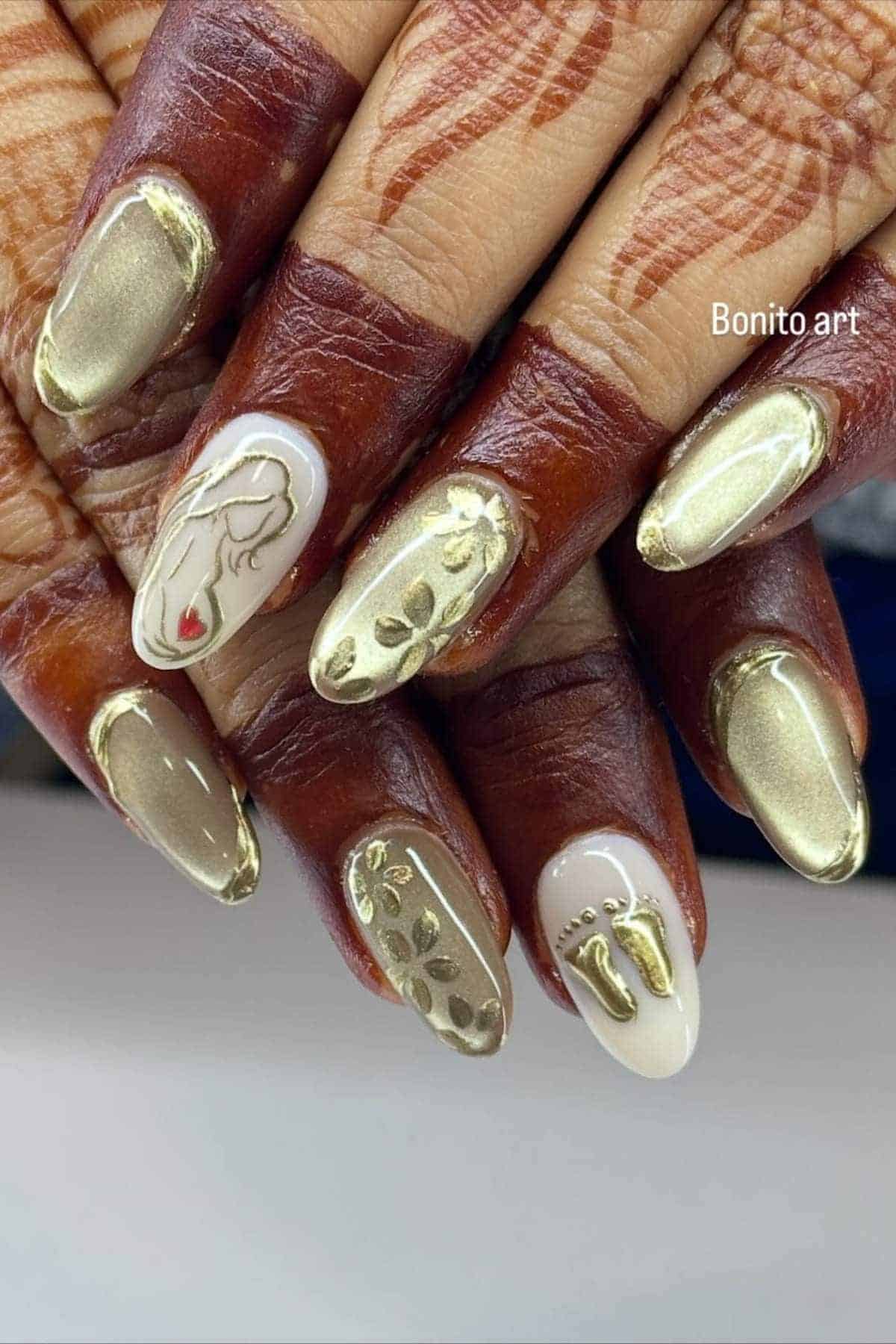 Close-up of hands with gold and white manicured nails featuring metallic accents, floral patterns, a face outline, and tiny foot designs; henna is visible on the fingers.