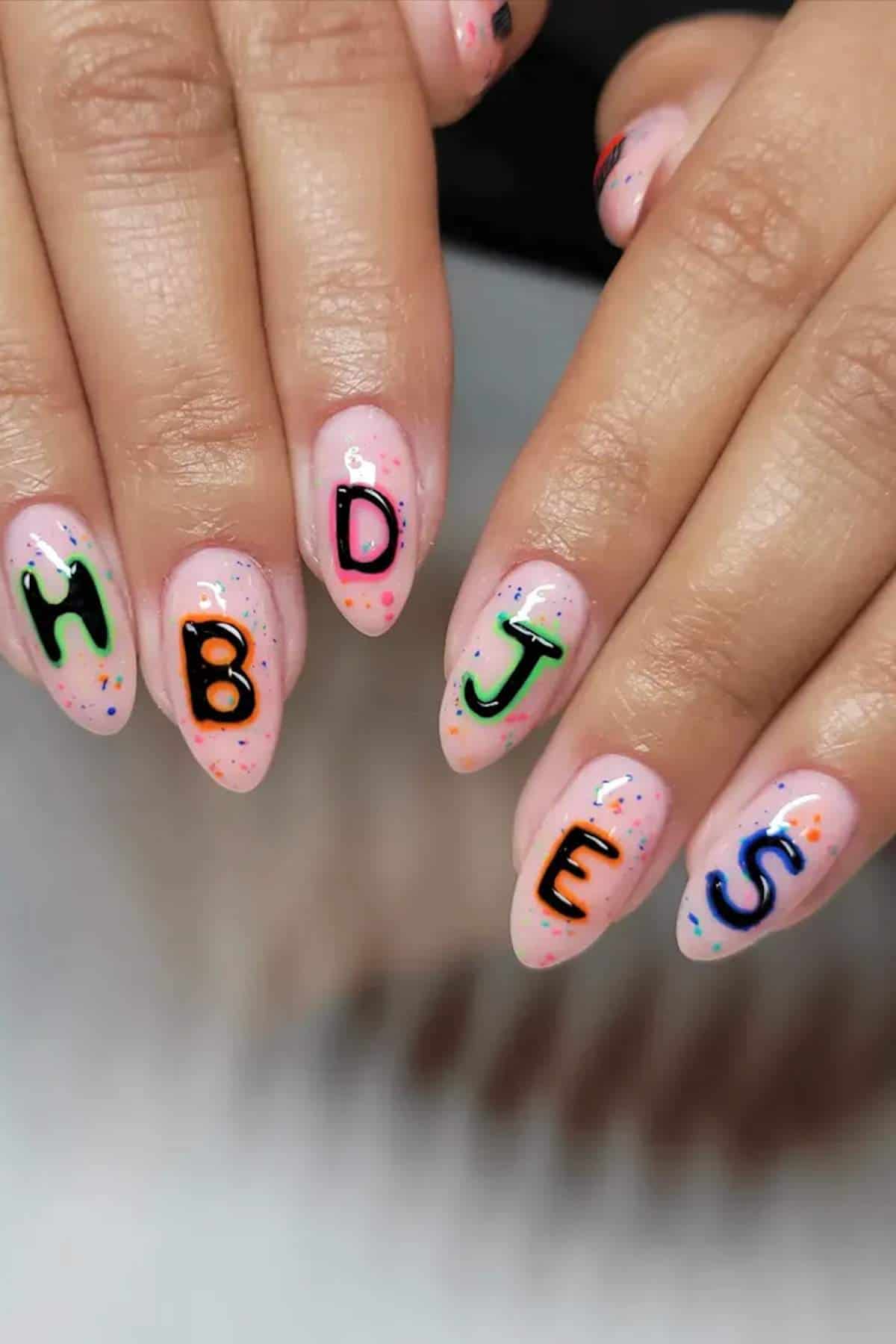 Close-up of hands with manicured nails featuring colorful letter designs: H, B, D, J, E, S on a light pink base with multicolored speckles.