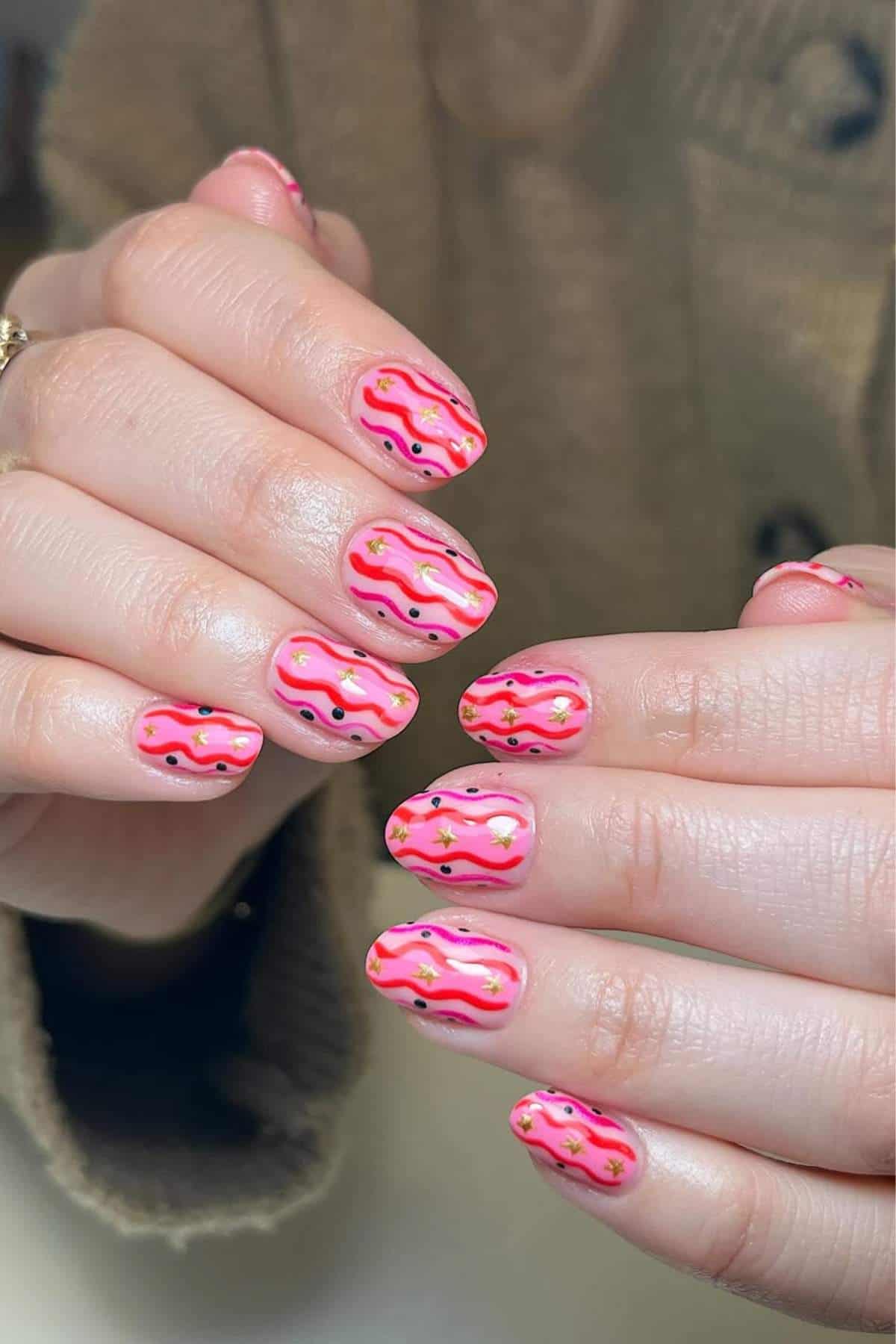 Close-up of hands with pink-themed nails featuring wavy lines in pink and orange, black dots, and small gold accents. The person is wearing a brown knit sweater.