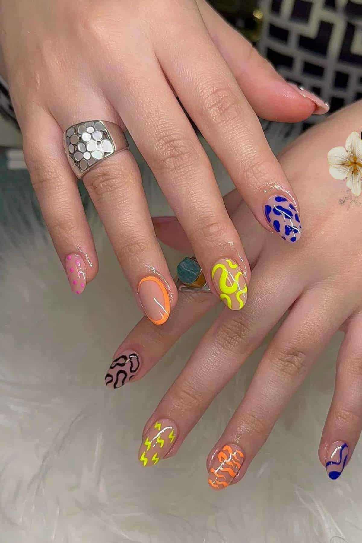 Hands with almond-shaped nails featuring multicolored abstract line designs, including pink, yellow, blue, orange, and black patterns. Hands are adorned with silver rings.