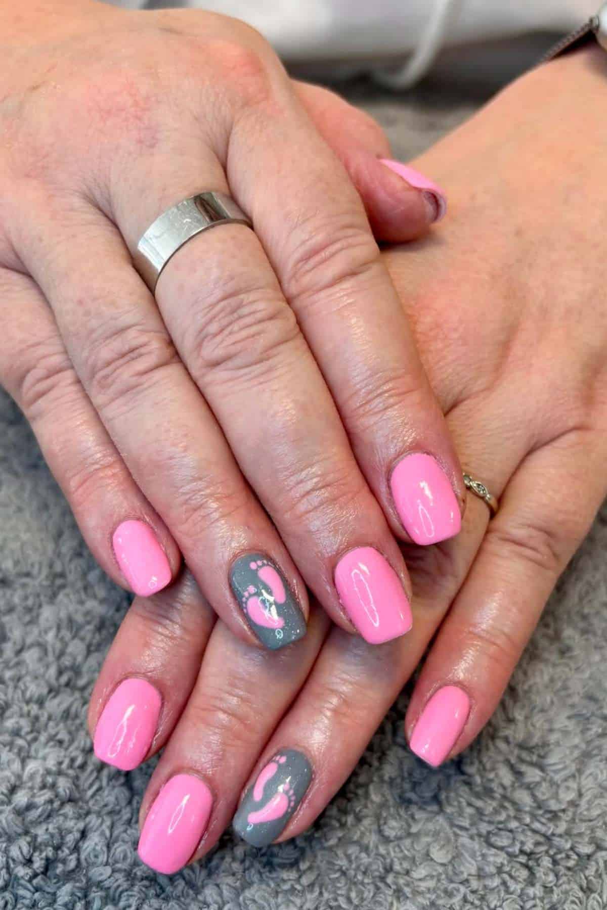 Hands with short, oval-shaped fingernails painted pink; ring fingernails are gray with white and pink ribbon designs. Hands are resting on a textured gray surface.