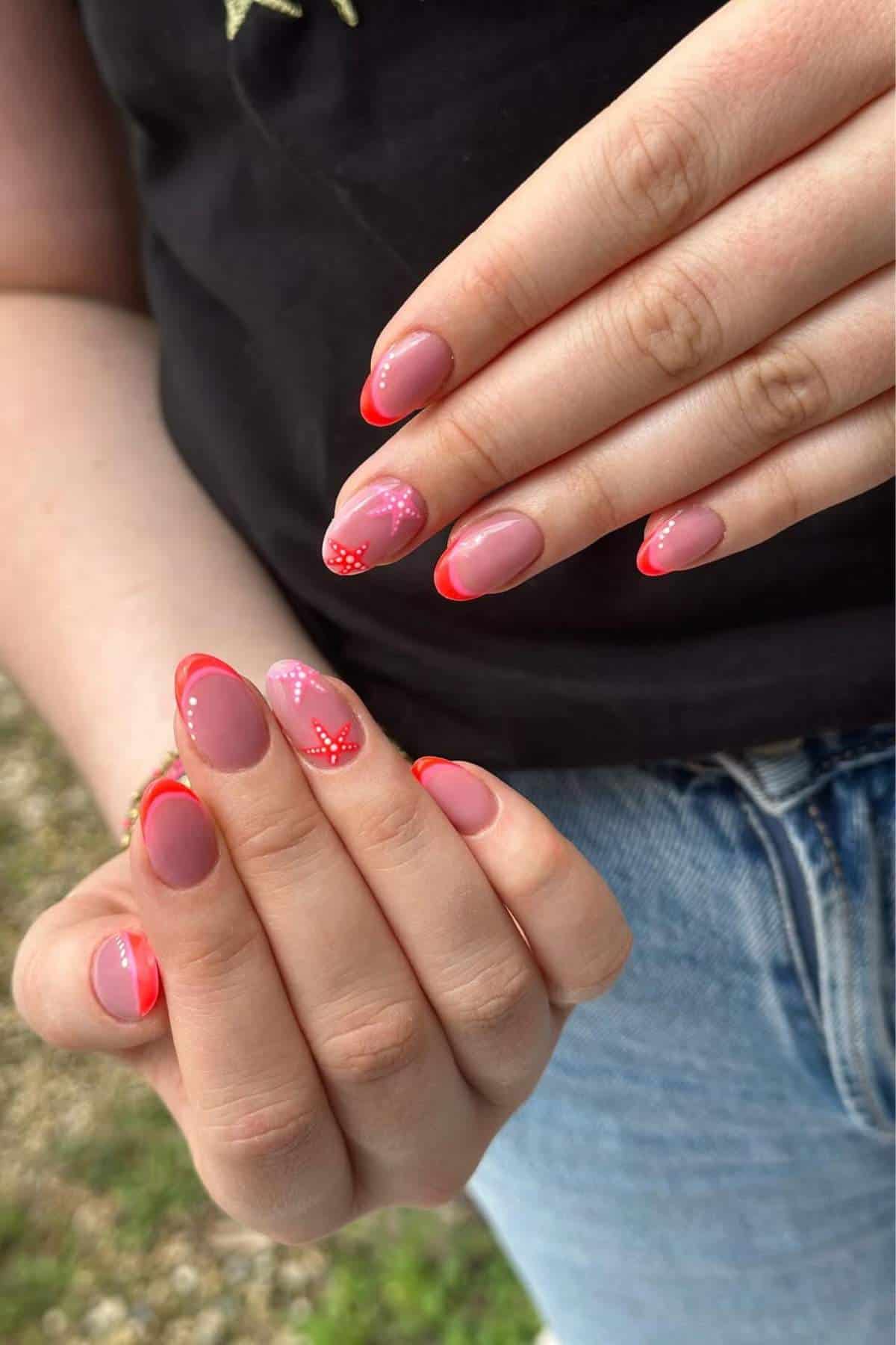 A person with oval-shaped nails painted pink with red French tips, dots, and heart designs, wearing a black top and blue jeans.