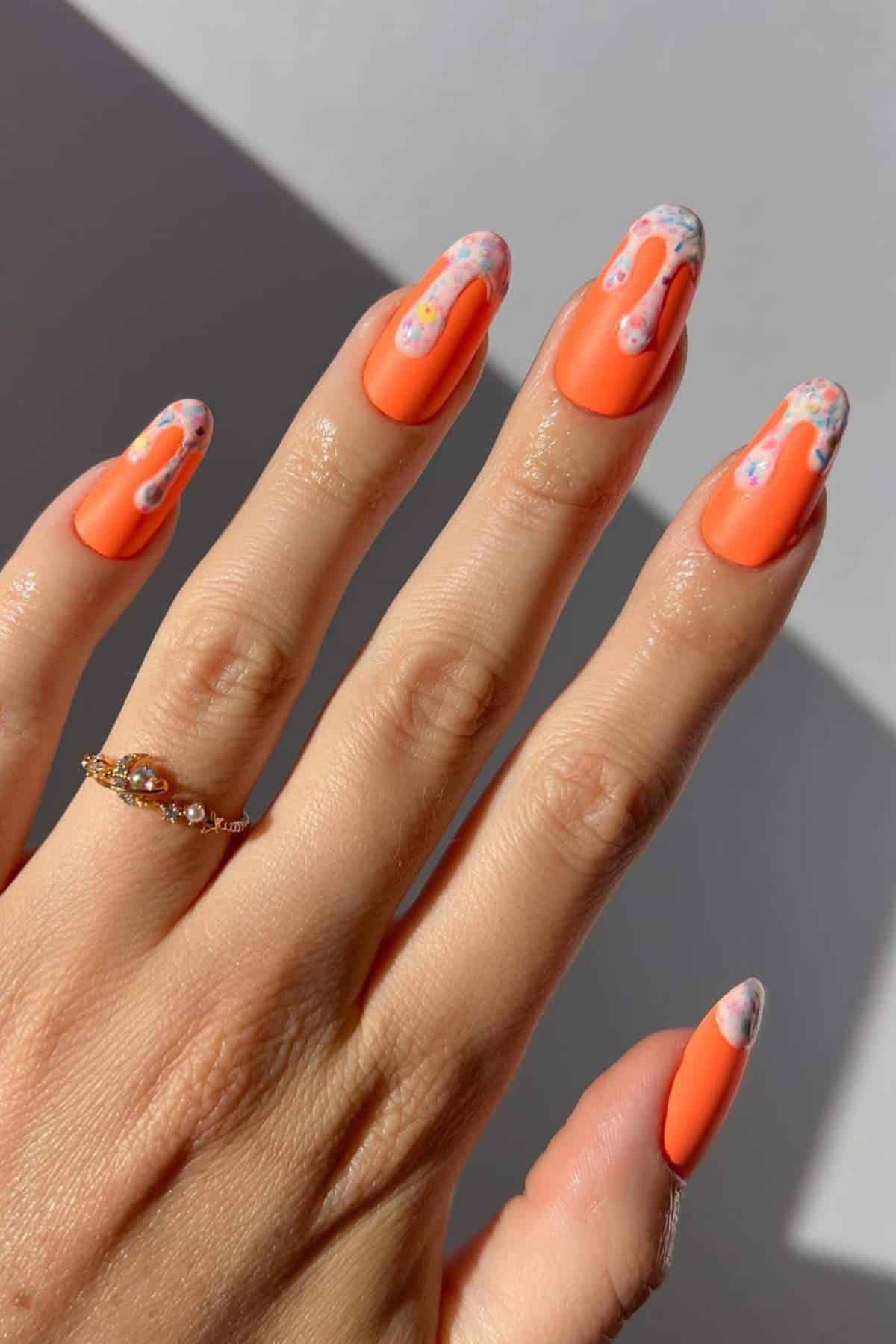 A hand with long, almond-shaped nails painted orange, featuring white tips with floral designs. The person is wearing a gold ring with a small stone.