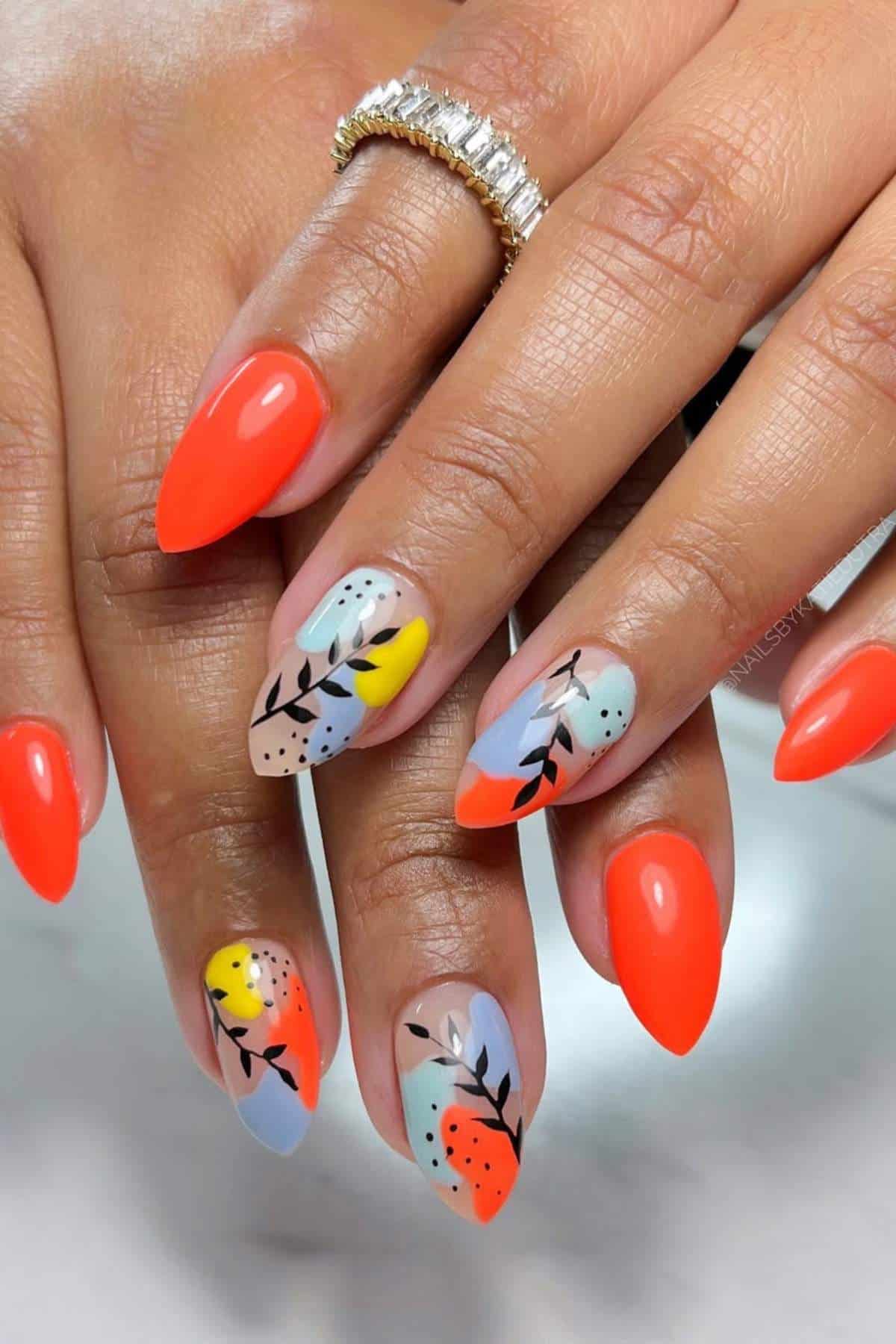 Hands with almond-shaped nails painted in bright orange and pastel colors with abstract black leaf designs and dotted patterns. A diamond ring is visible on one finger.