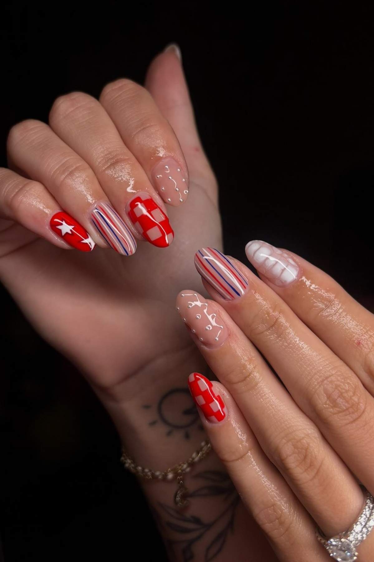 A close-up of hands with almond-shaped nails featuring various red, white, and nude designs, including stars, checkers, stripes, and abstract dots.