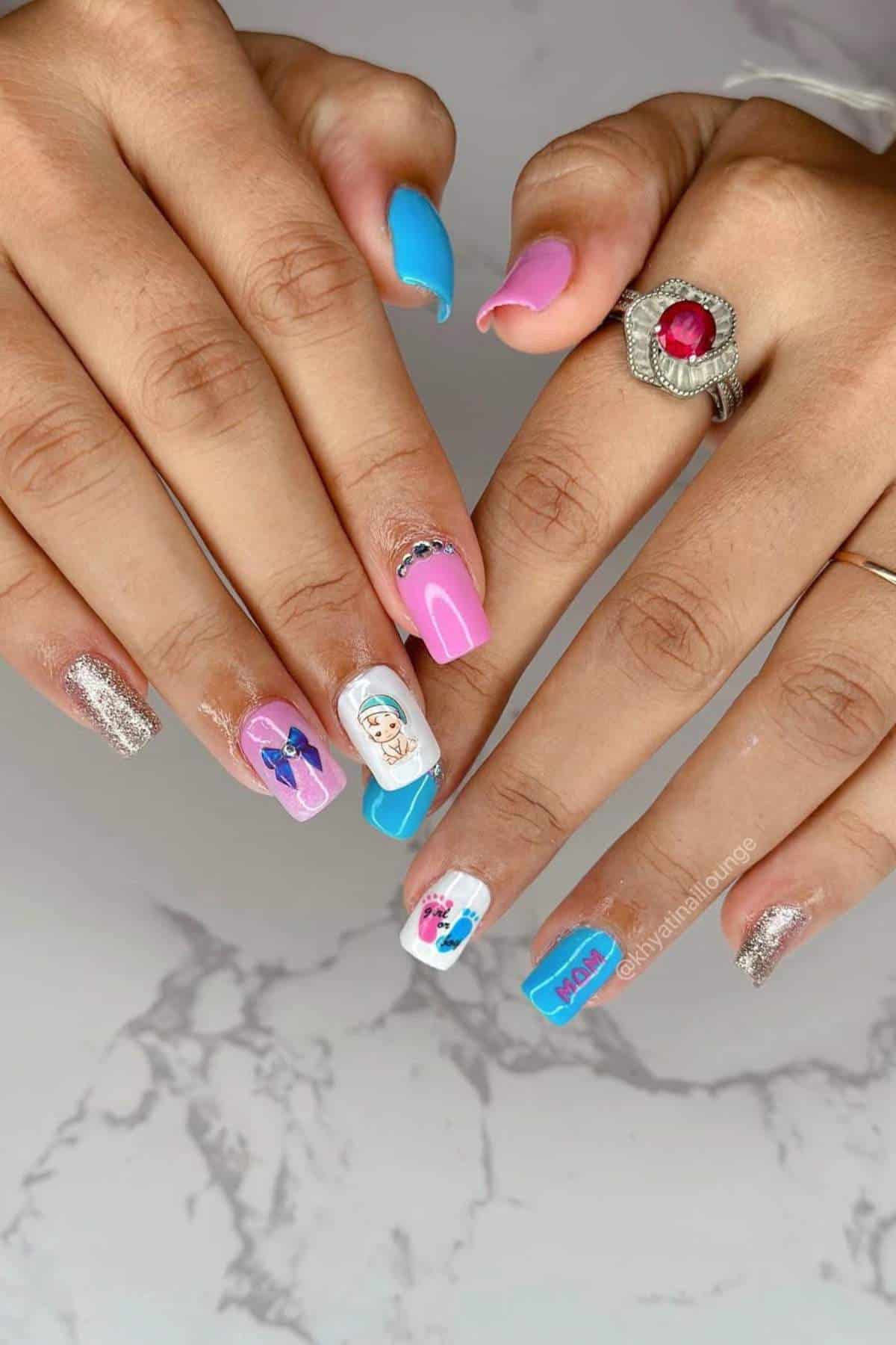 Hands with manicured nails featuring blue, pink, white, and silver designs, including cartoon faces, bows, and "Mom" text; a ring with a red stone is worn on one finger.