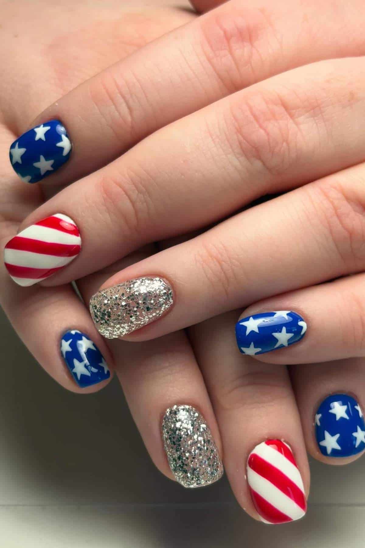 Close-up of hands with manicured nails featuring American flag designs, blue with white stars, red and white stripes, and silver glitter accents.