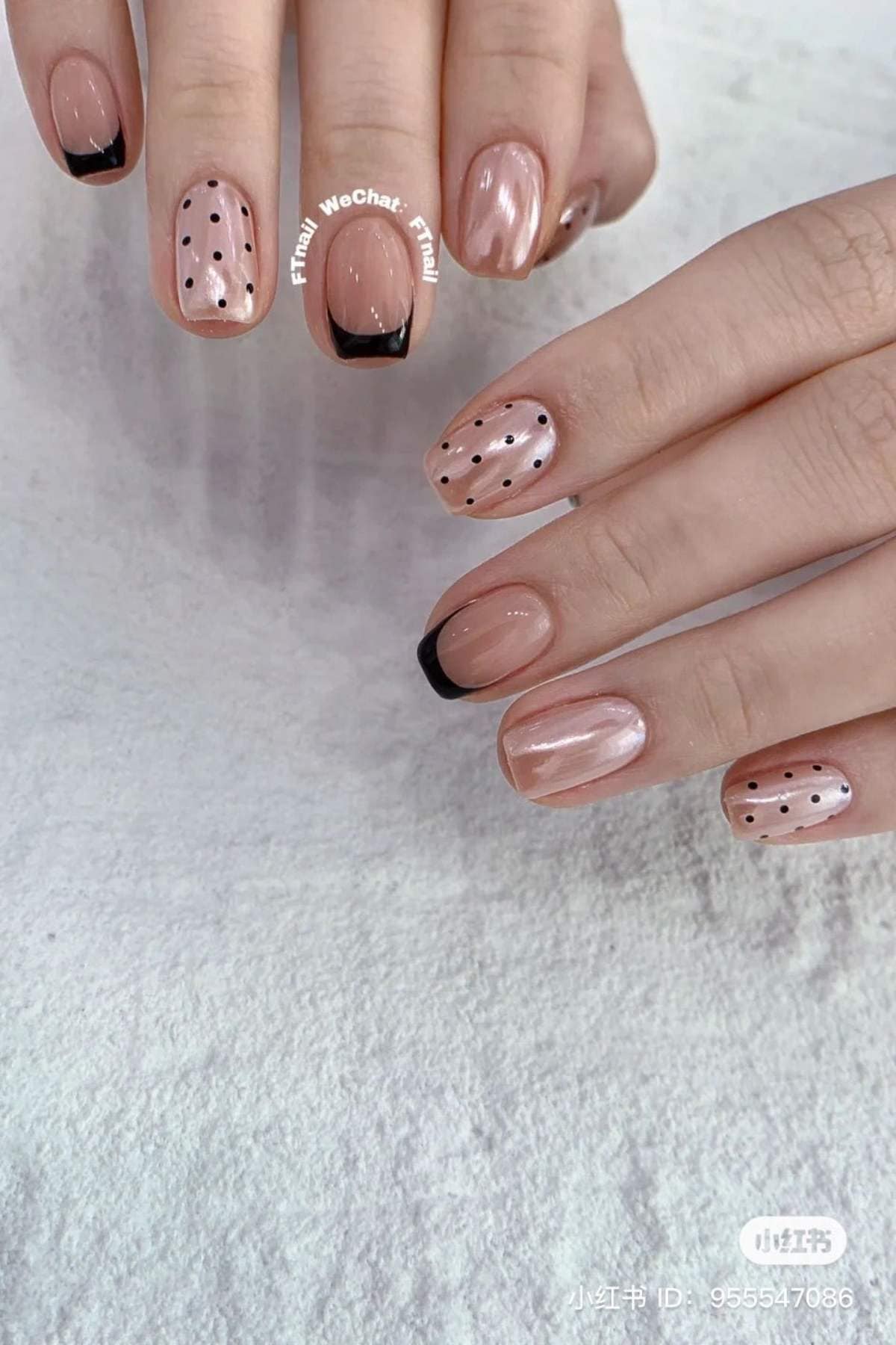 Hands with glossy nude-pink manicured nails featuring black French tips and black polka dot designs, shown on a white textured surface.