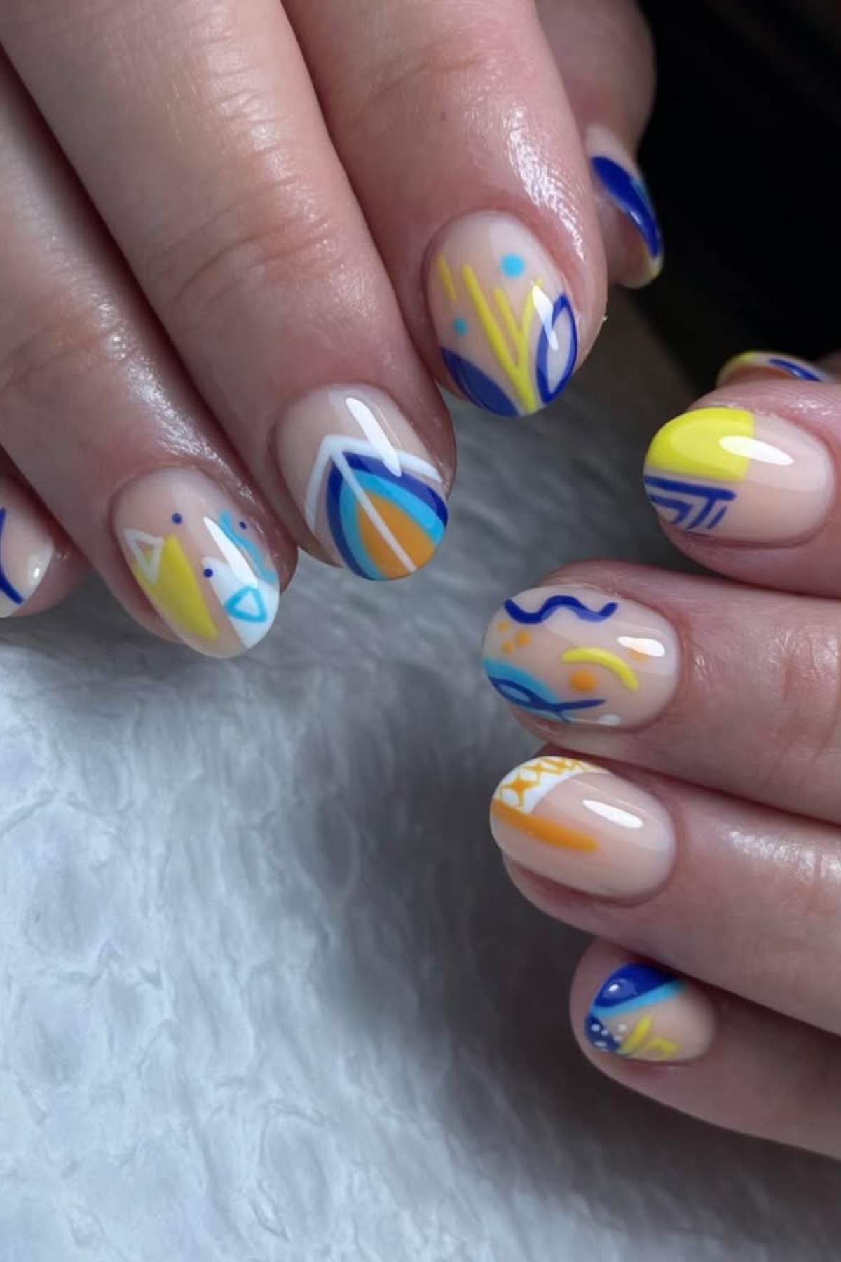 Hands with short, oval-shaped nails featuring abstract designs in blue, yellow, and white on a neutral base.