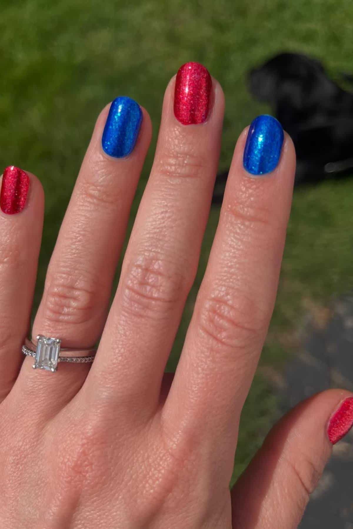 A hand with red and blue glitter nail polish, wearing a rectangular diamond ring, is shown outdoors with grass in the background.