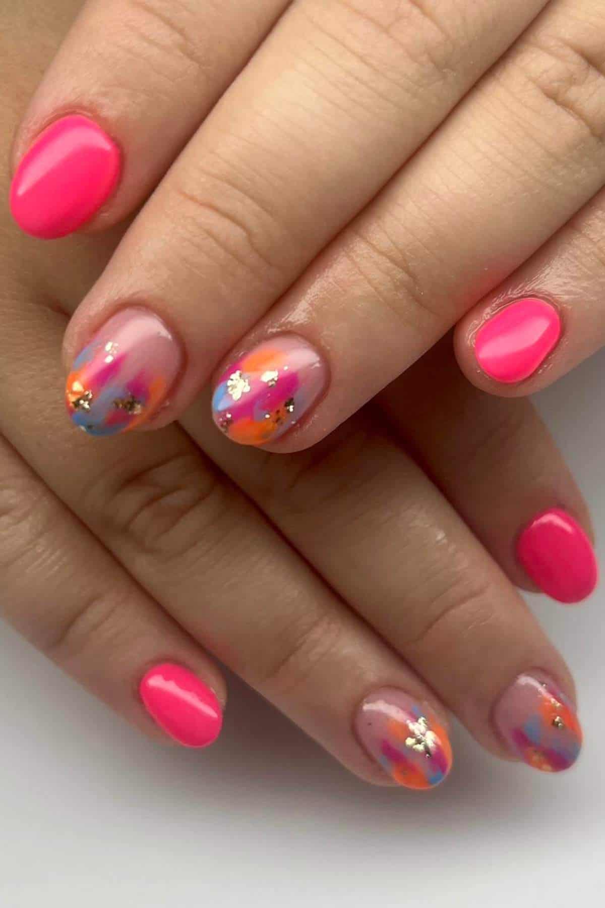 Hands with short, oval-shaped nails featuring bright pink polish and accent nails decorated with abstract colorful designs and gold foil on a nude base.