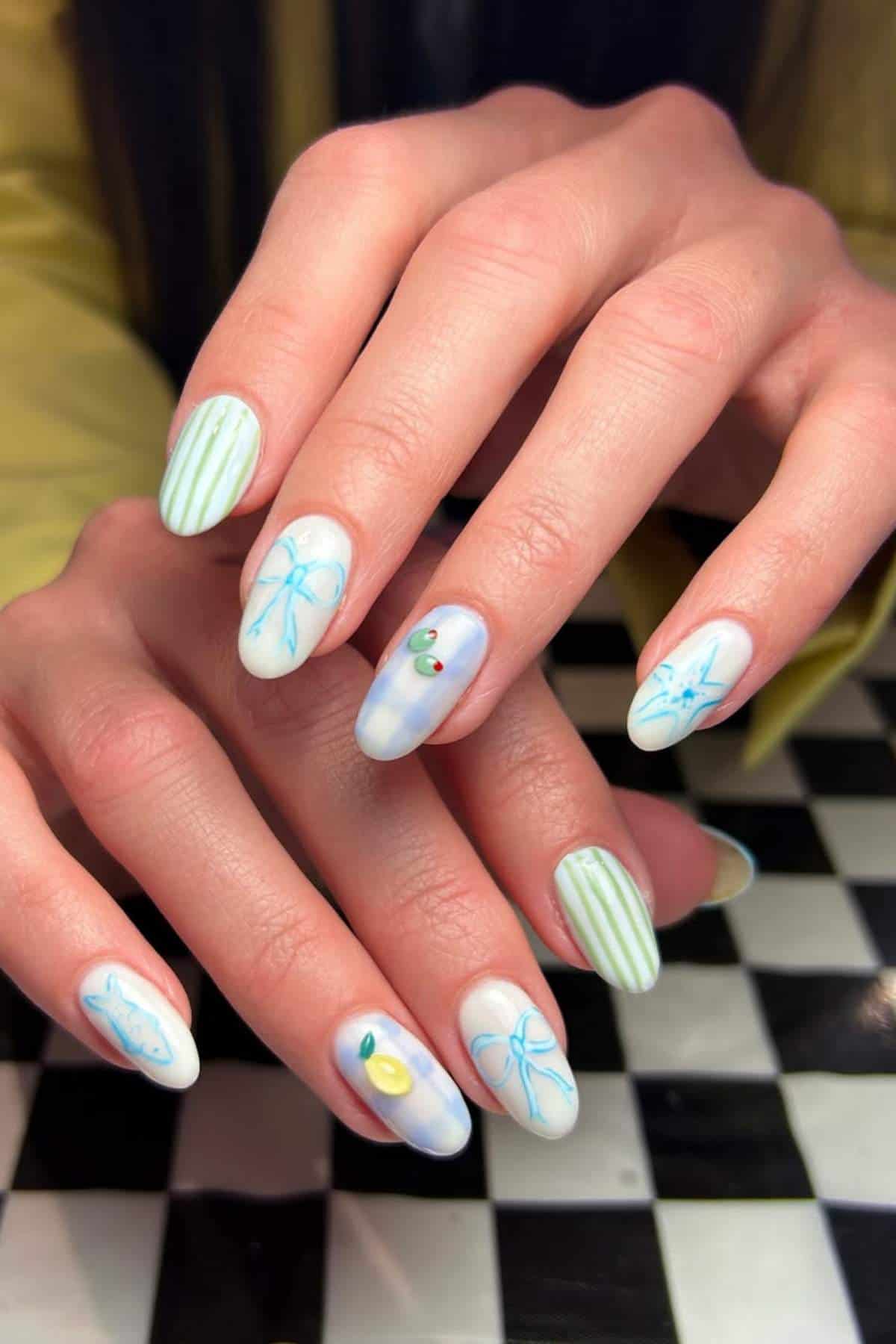 Hands with almond-shaped nails featuring pastel green stripes, blue abstract lines, and small 3D fruit decorations, displayed over a black-and-white checkered surface.