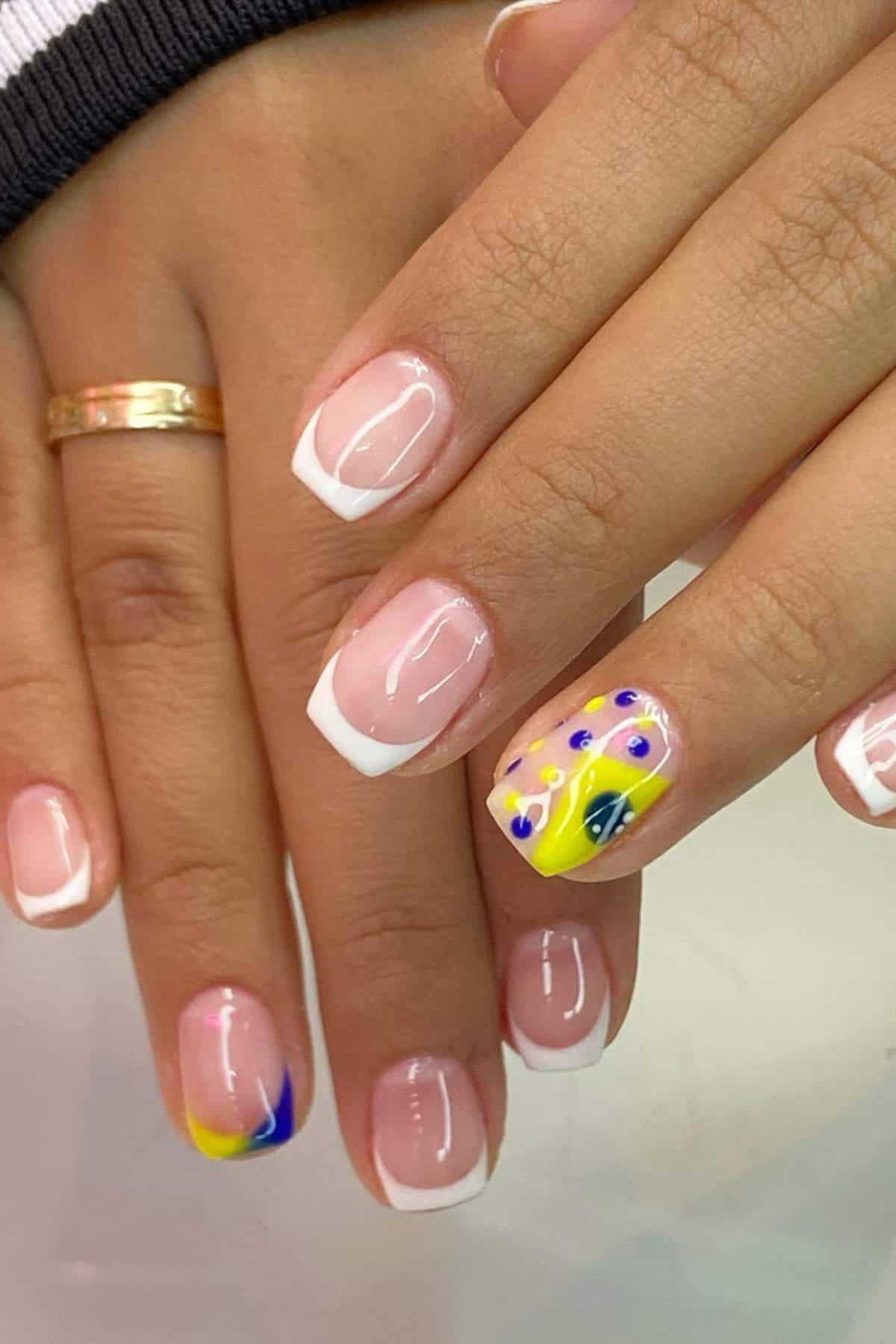 A hand with short, square nails featuring classic French tips and one accent nail with yellow, blue, and purple abstract dot and swirl designs.