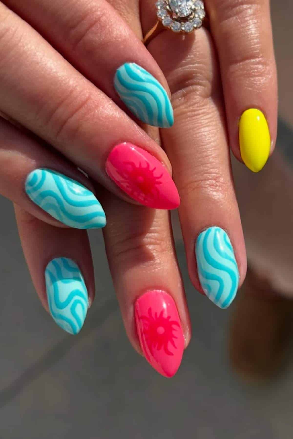 A hand with almond-shaped nails featuring blue wave patterns, two pink nails with sun designs, and one bright yellow nail.