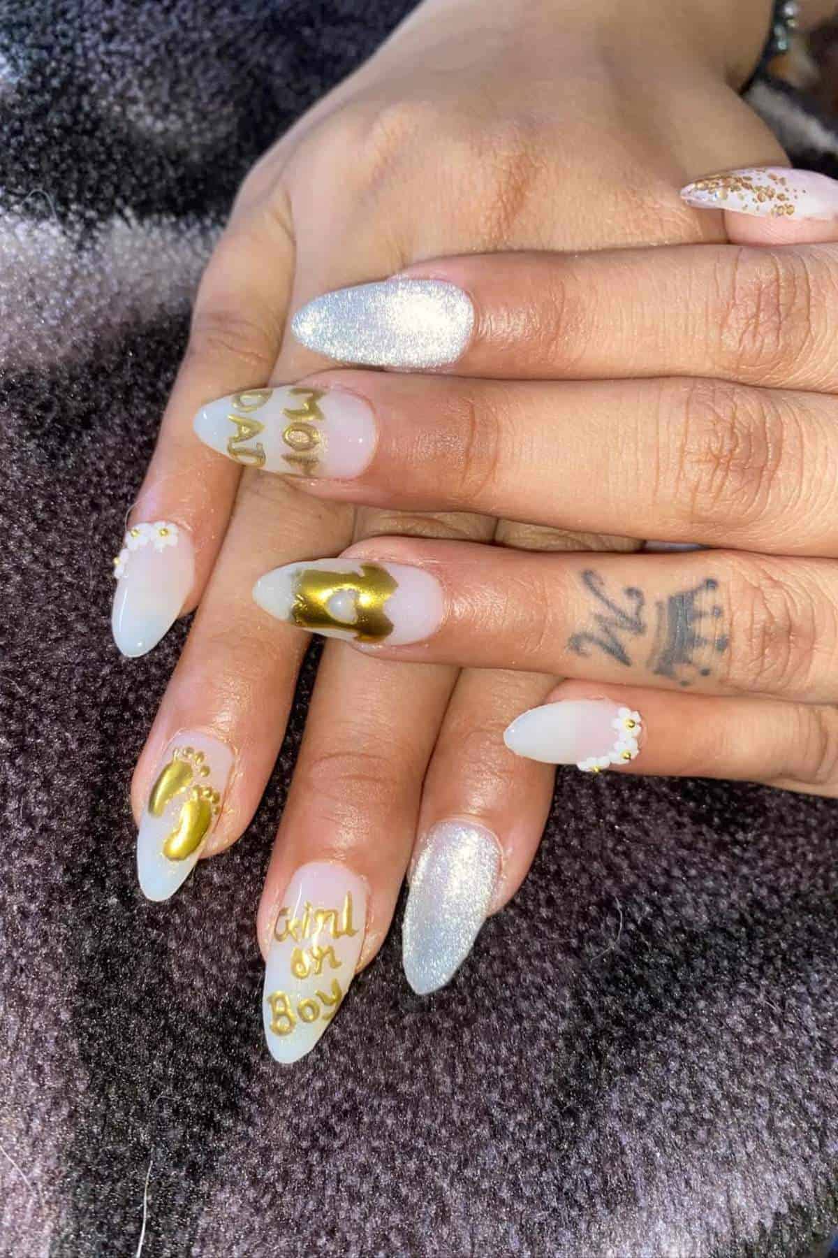 Two hands with long, almond-shaped nails featuring gold and silver designs, including “Boy or Girl” text and bows, resting on a patterned fabric.