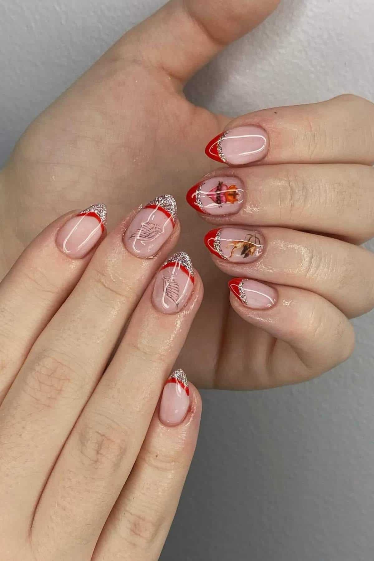 Hands with almond-shaped nails featuring red French tips, silver glitter accents, white leaf designs, and realistic floral nail art on two accent nails.